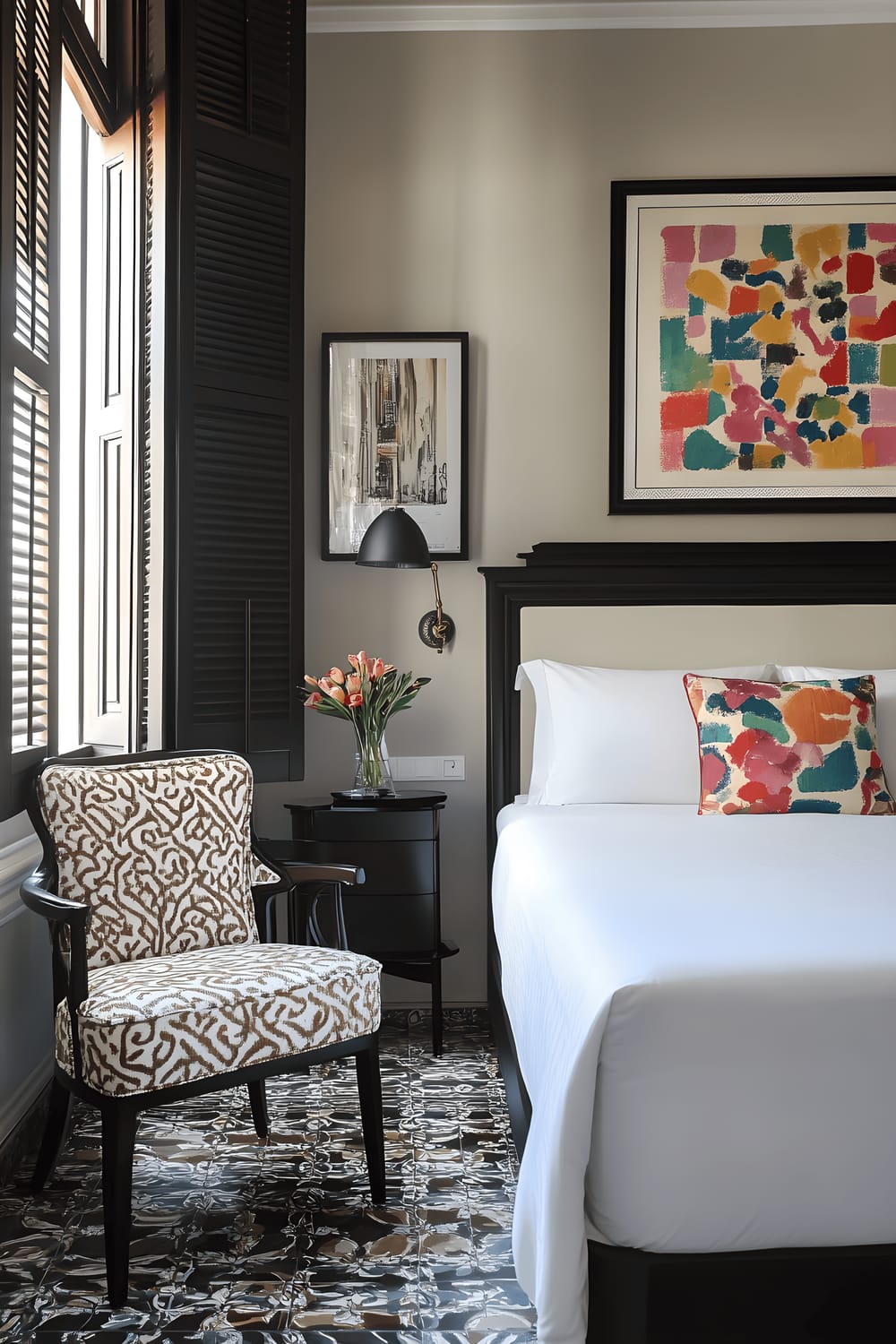 A front view of a stylish guest room, showcasing a queen-sized bed with a unique Catalan-designed headboard, dressed in white linens with colourful accent pillows. Adjacent to the bed is a sleek nightstand bearing a vintage-style lamp and a small vase of fresh flowers. The room also features vibrant, local artwork on its walls, and a comfortable, patterned armchair situated next to a small writing desk. The room is filled with natural light streaming in from large windows with decorative shutters.