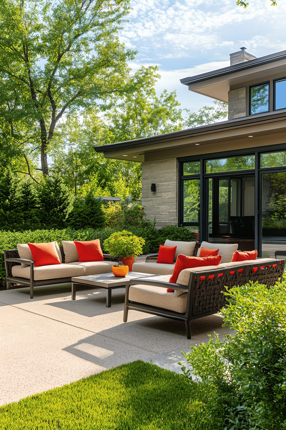 A well-kept backyard with a clean, simple patio, complemented by beige outdoor seating with vibrant red and orange cushions. The patio is surrounded by green shrubbery and overlooks a lawn. A colorful potted plant graces the concrete patio while large house windows in the background enhance the indoor-outdoor connection.