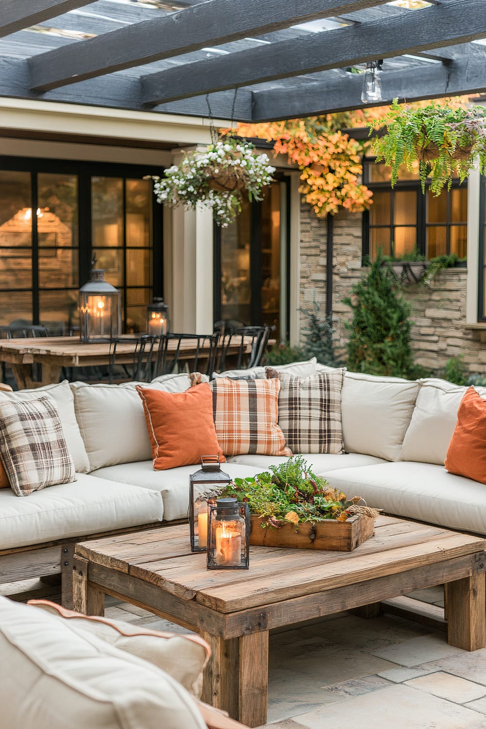 An outdoor seating area featuring a beige sectional sofa adorned with plaid and orange cushions. A wooden coffee table in the center holds two lanterns with lit candles and a wooden crate filled with potted plants. Hanging plants are suspended from the overhead pergola, and a larger dining table with chairs and lanterns can be seen in the background. Stone accents and ivy climbing the walls add to the aesthetic of the cozy outdoor space.