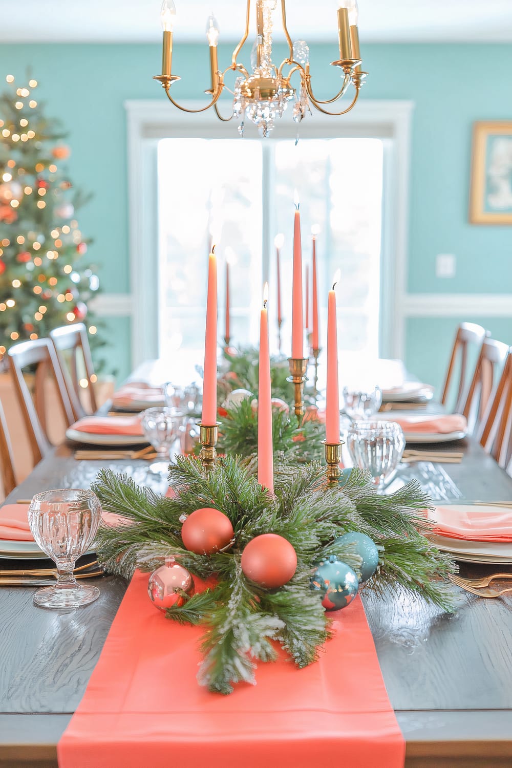 Elegant holiday dining table with a festive centerpiece featuring pink candles, greenery, and ornaments. The table is set with pink napkins, white plates, and brass candle holders. A Christmas tree is adorned with lights and decorations in the background, creating a warm and inviting ambiance under a gold chandelier.