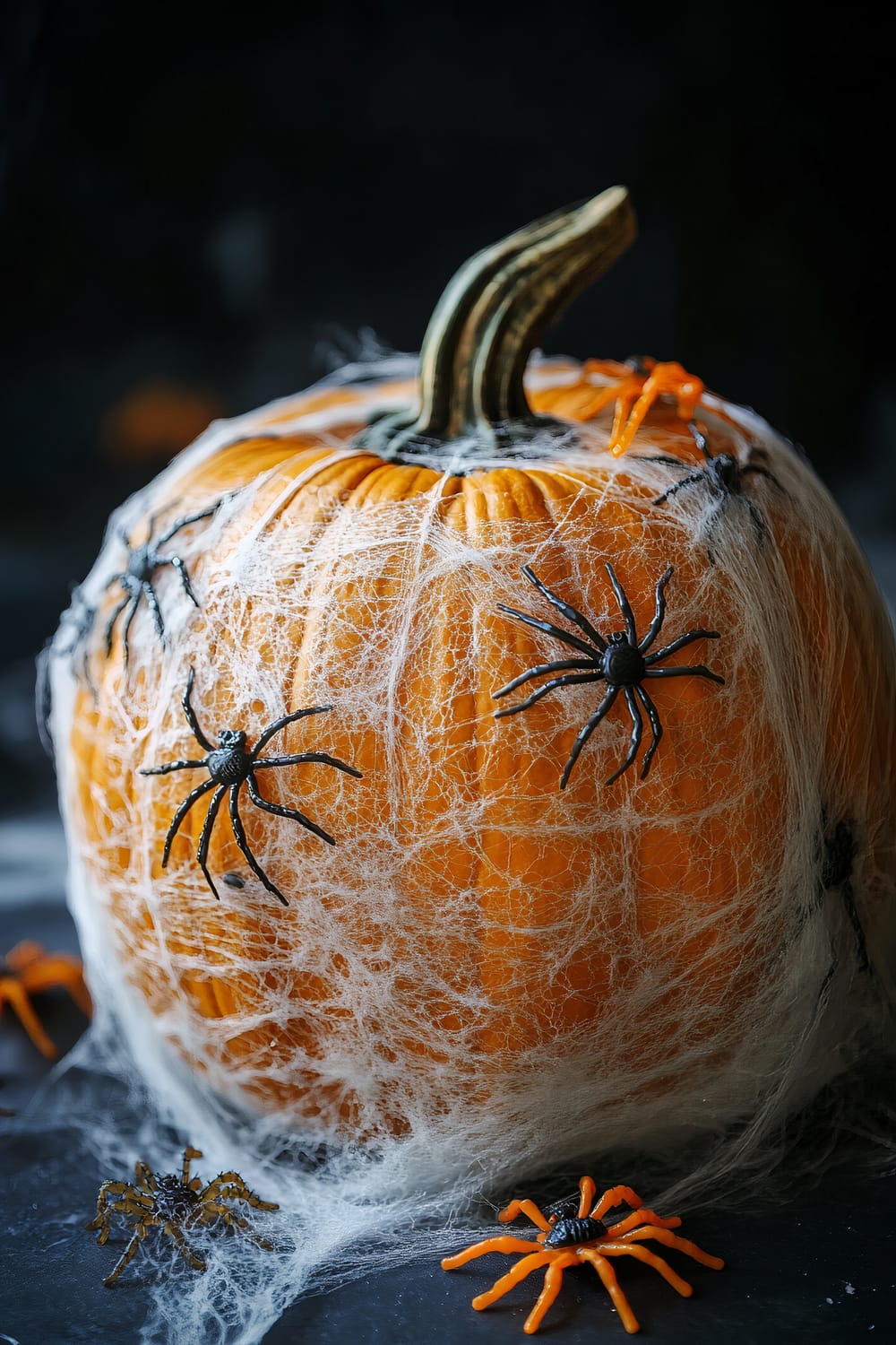 A close-up of an orange pumpkin decorated for Halloween. The pumpkin is covered in synthetic spiderwebs and adorned with black and orange plastic spiders. The background is dark, emphasizing the Halloween theme.