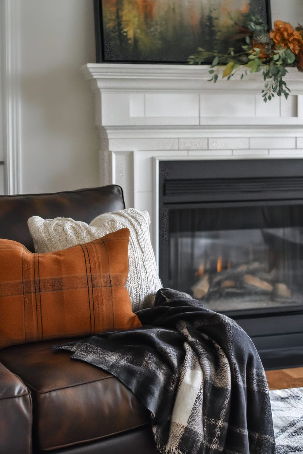 A close-up of a living room interior, showcasing a warm and inviting arrangement. A brown leather sofa is adorned with two cushions, one a cozy white knit and the other a plaid-patterned orange fabric. A black and white plaid throw is casually draped over the arm of the sofa. In the background, a modern white mantelpiece frames a gas fireplace with burning logs. Above the mantel, a painting of trees in autumnal hues adds a touch of nature to the decor. A simple fall-themed floral arrangement is also placed on the mantel.