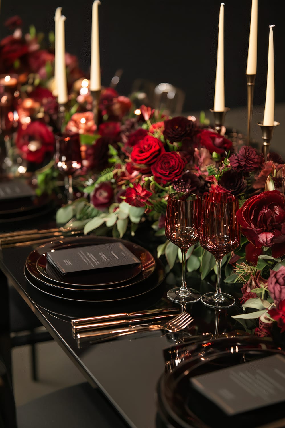 A sophisticated dining table set with deep red floral arrangements and dark tableware. Tall, slender candles in brass holders are lit, casting a warm glow over the setting. Two red wine glasses sit next to black plates with elegant, printed menus. Cutlery is precisely placed to the right of the plates, and the overall ambiance is luxurious and romantic.