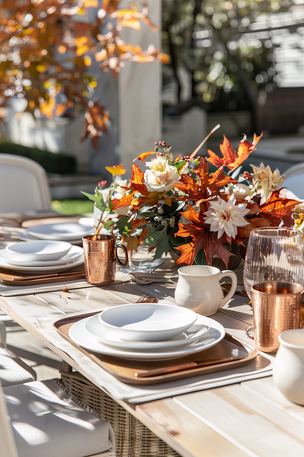 An outdoor dining table set for fall, featuring a central arrangement of autumn foliage and flowers in red, orange, and white tones. The table has white plates placed on wooden chargers, and copper mugs and cutlery enhancing the seasonal theme. Soft sunlight filters through the trees, casting a dappled shadow on the table.