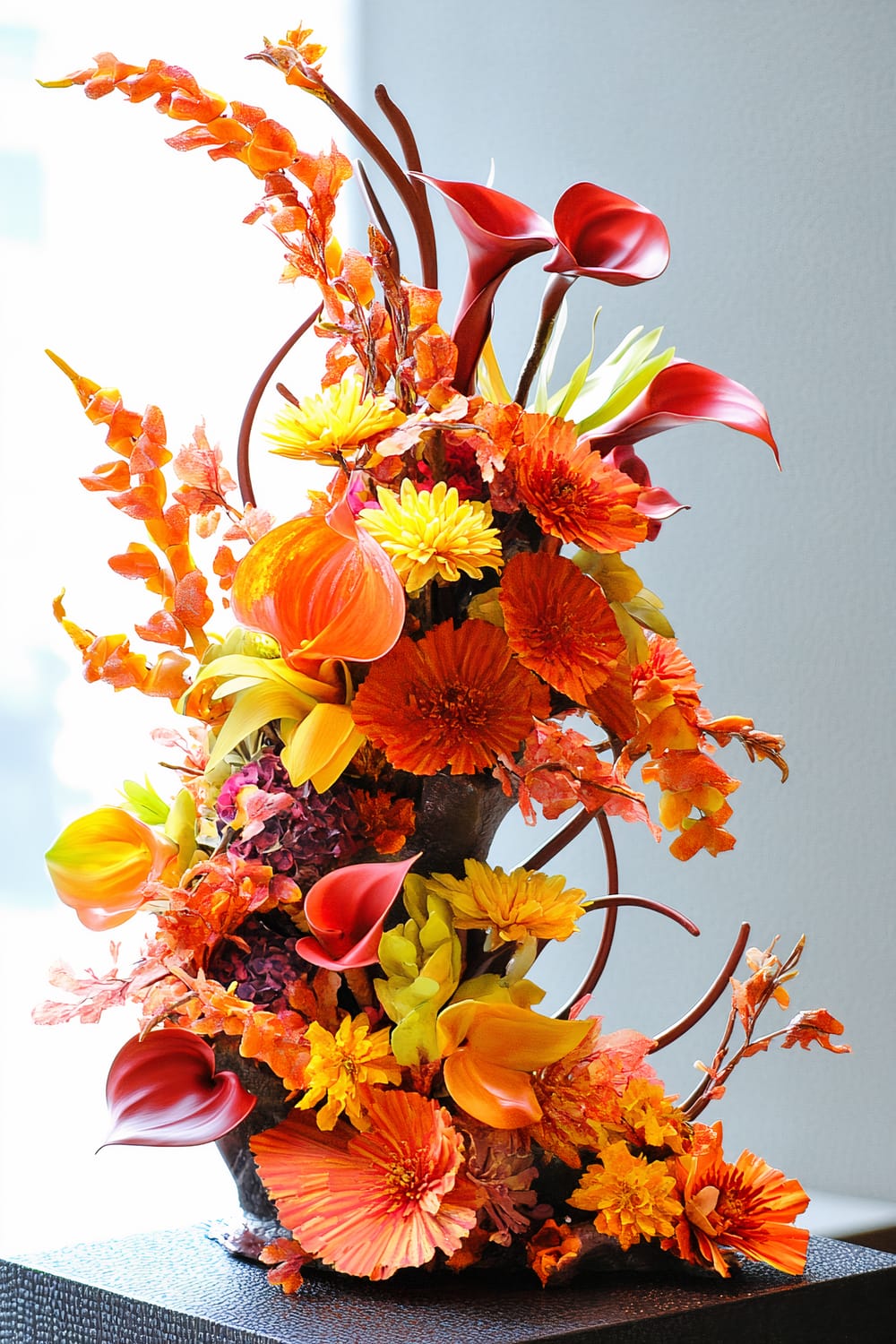 A vibrant floral arrangement featuring a mix of red, orange, yellow, and green flowers. The flowers include calla lilies, marigolds, and various other blooms, gracefully arranged in a dynamic and cascading fashion. The arrangement is set against a neutral background.