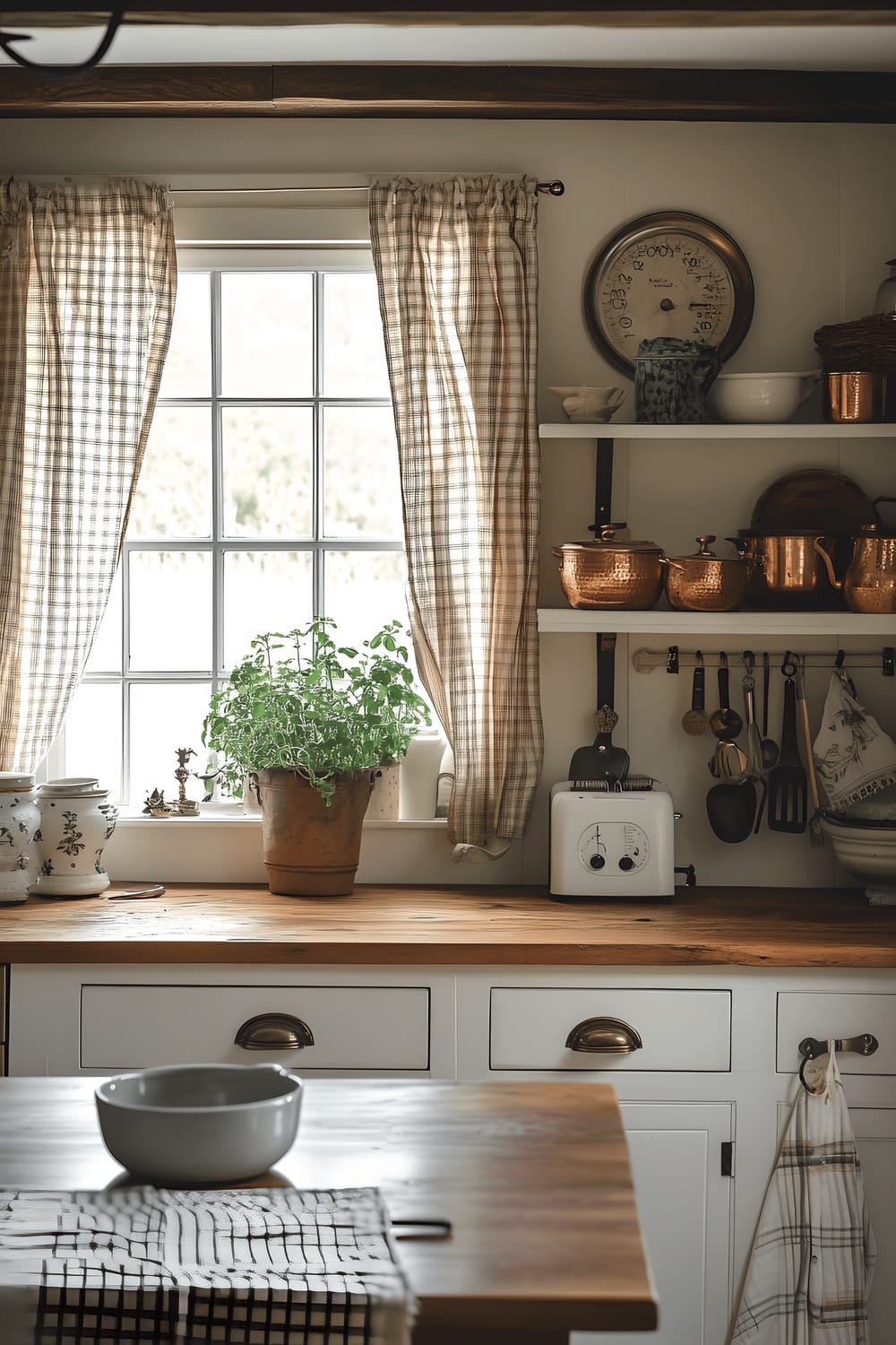 A rustic French Country kitchen with distressed white cabinetry, warm wooden countertops adorned with copper pots and vintage kitchenware, a wood table set with linen and a vase of wildflowers, and a large window that lets in natural light from an herb garden. The room is illuminated by a wrought iron chandelier and features open shelves with rustic kitchenware, exposed wooden beams, and plaid curtains.