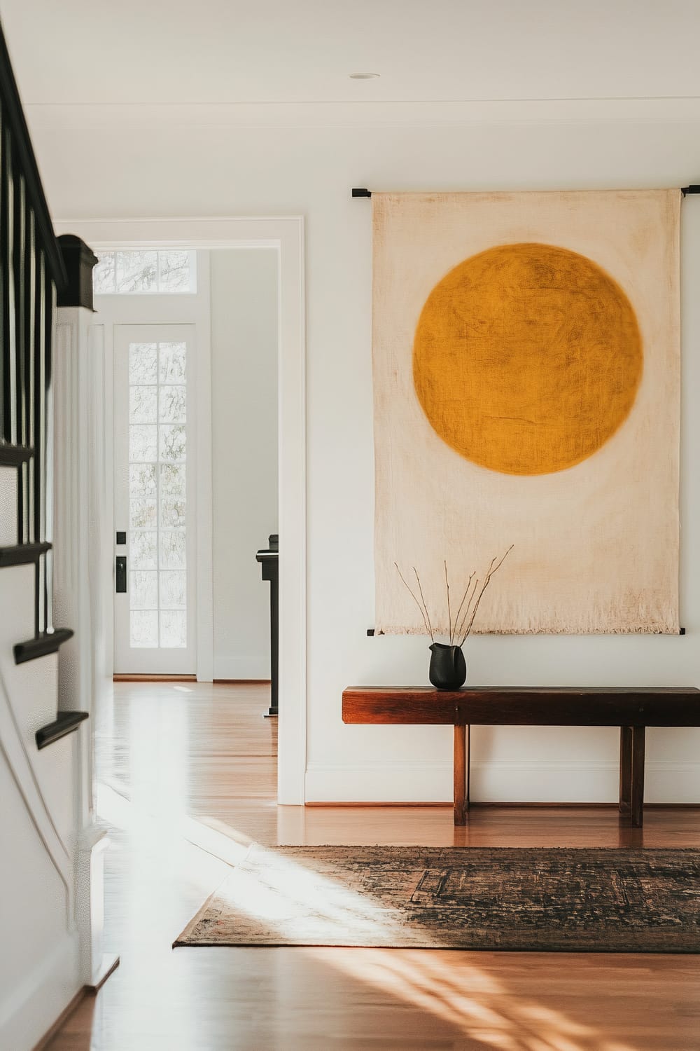 A minimalist entryway with a focus on natural light and simple, organic elements. The space features a wooden bench with a black vase holding a few branches. Above the bench hangs a large tapestry with a prominent yellow circle. The flooring is light wood, and a rug with a subtle pattern lies in front of the bench. The view extends to a well-lit room with glass doors and a glimpse of a staircase to the left.