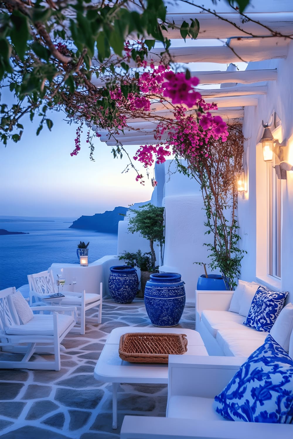 A stunning and picturesque patio styled in themes reminiscent of Santorini. The patio features white furniture accented with deep cobalt blue cushions and floral patterns, and blue ceramic planters holding bougainvillea trees. The trees climb and spread over a white pergola, adding natural beauty and providing shade. The floor is an uneven but beautiful spread of whitewashed stone, giving off a rustic feel. The vast blue Aegean Sea can be seen in the background, painted vibrantly by the sunset.