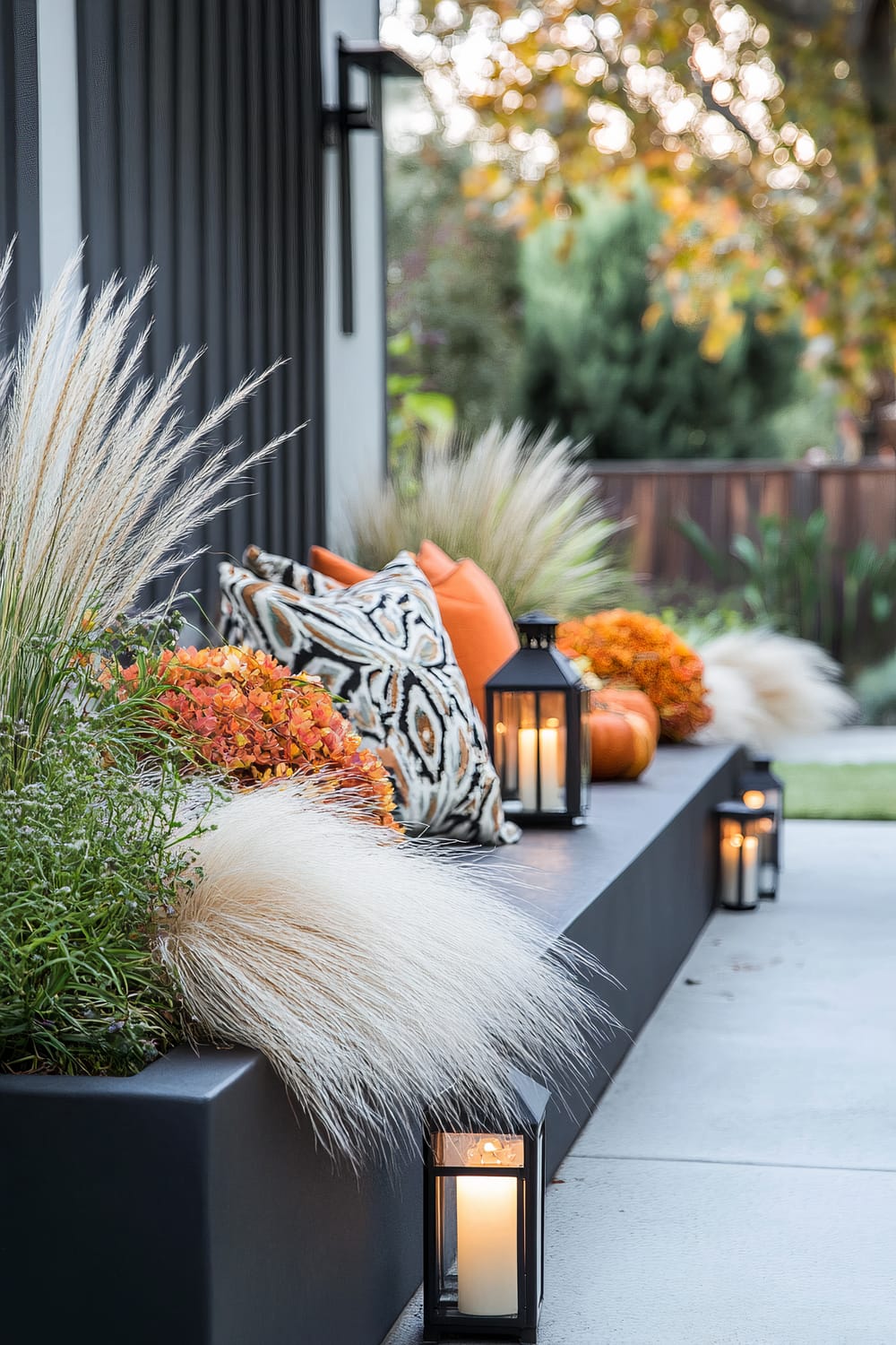 An elegant outdoor seating arrangement showcases a modern design with a touch of cozy charm, perfect for an autumn setting. The seating area is adorned with a collection of patterned and solid orange cushions, reflecting a warm palette. Potted ornamental grasses, orange chrysanthemums, and pumpkins add to the fall theme. Black lanterns with lit white candles are strategically placed along the edges, adding a soft, inviting glow. The backdrop features a neatly maintained garden with lush greenery and a wooden fence in the distance.