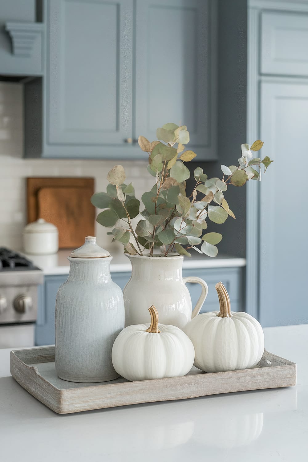 A minimalist kitchen counter is adorned with a decorative wooden tray displaying autumn-themed items. The tray holds two white pumpkins, a ceramic jar with a lid, and a ceramic pitcher with eucalyptus branches. The background features soft blue cabinetry with subtle molding, a gas stove, and white subway tile backsplash.