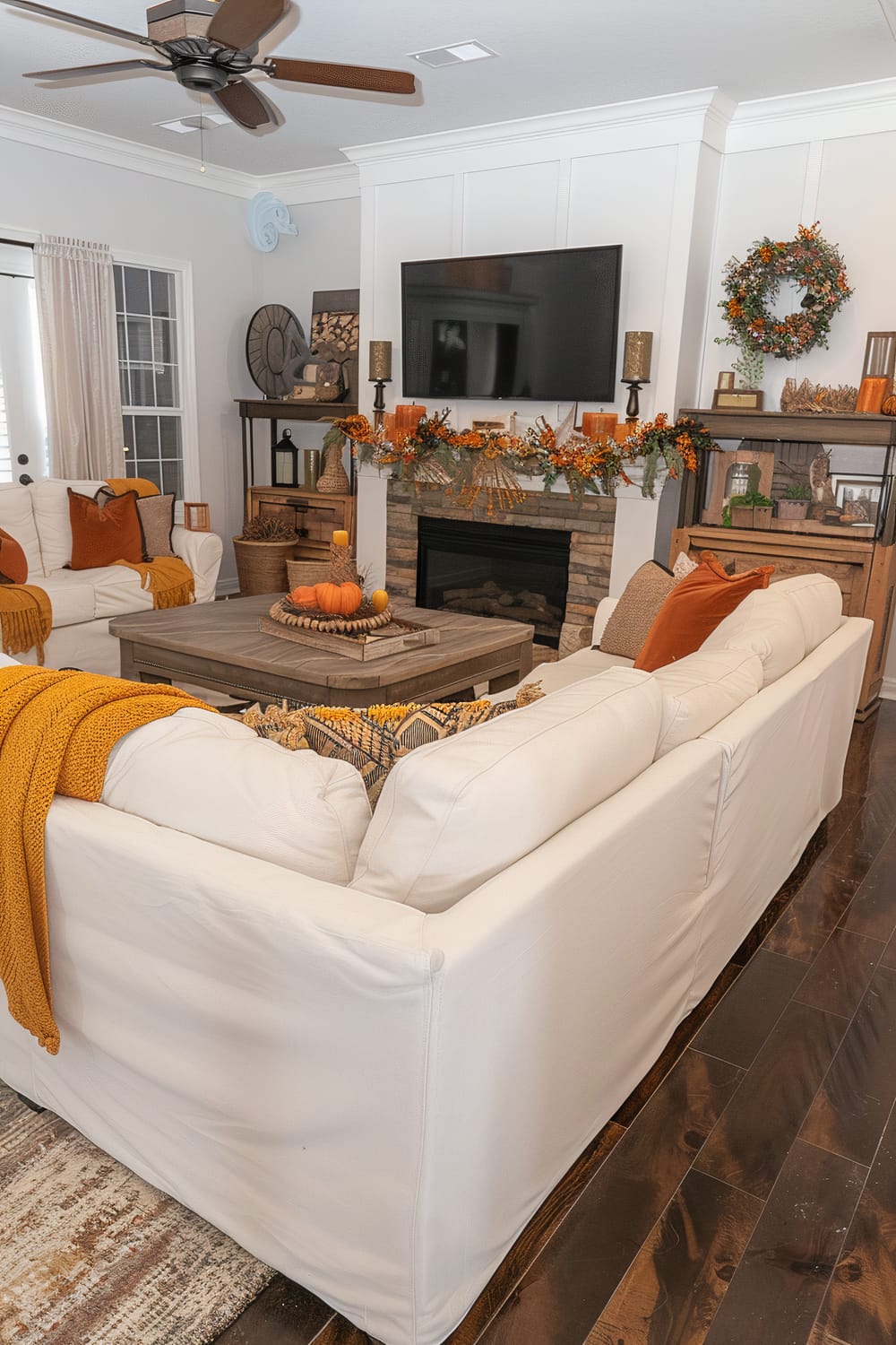 This living room features a cozy and warm autumn-themed decor. The white sofas are accessorized with orange, mustard, and patterned throw pillows, along with blankets that match the autumnal color scheme. A rustic wooden coffee table in the center is adorned with pumpkins and candles. A stone fireplace with a festive garland and candles serves as the focal point, and a mounted flat-screen TV is above it. Shelves display seasonal decorations, including a wreath and various trinkets. A ceiling fan and large windows complete the inviting space.