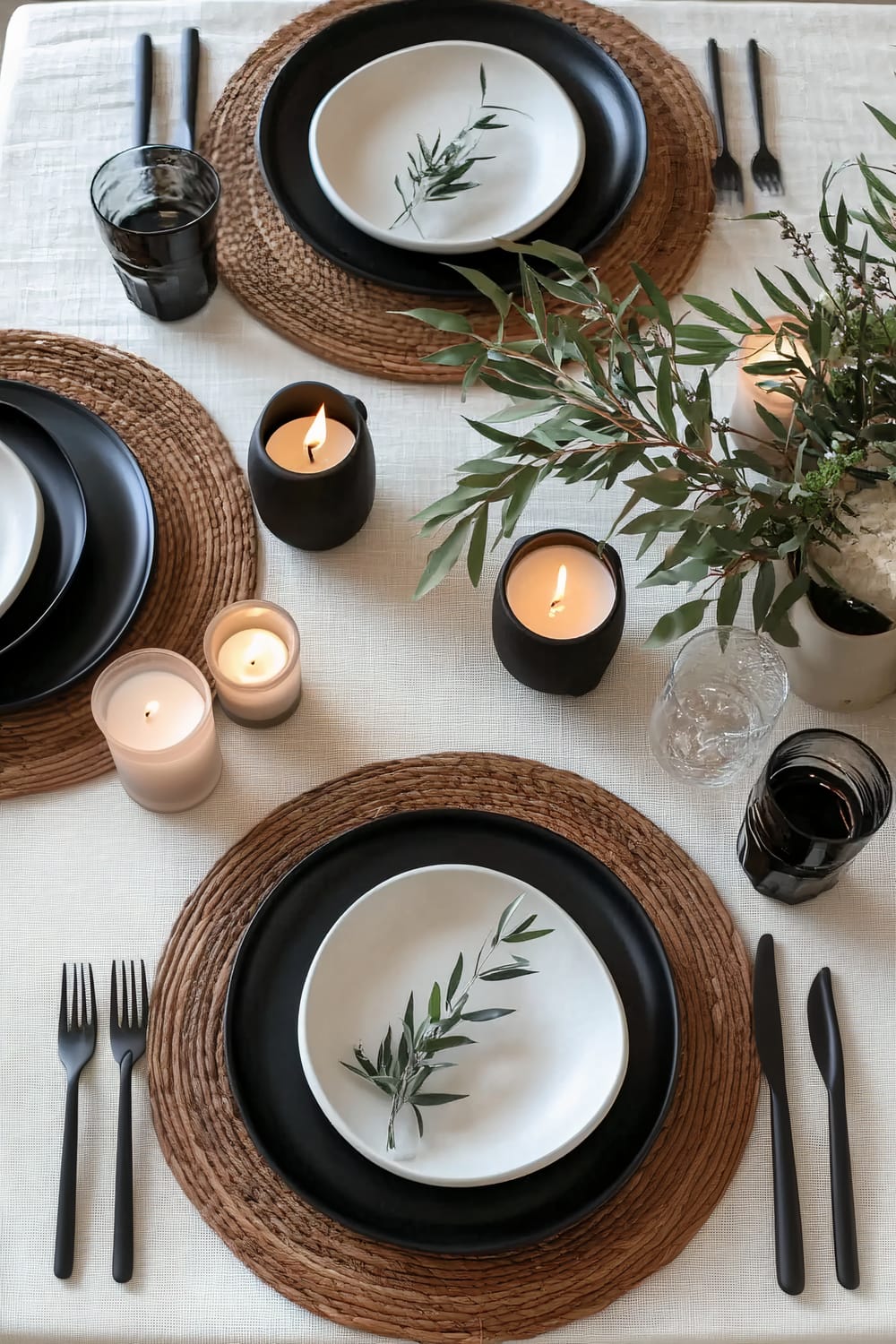 Modern Japandi-inspired Thanksgiving table setting featuring sleek black and white tableware atop natural wood chargers. A minimalist centerpiece of greenery and scattered candles completes the elegant, serene atmosphere.