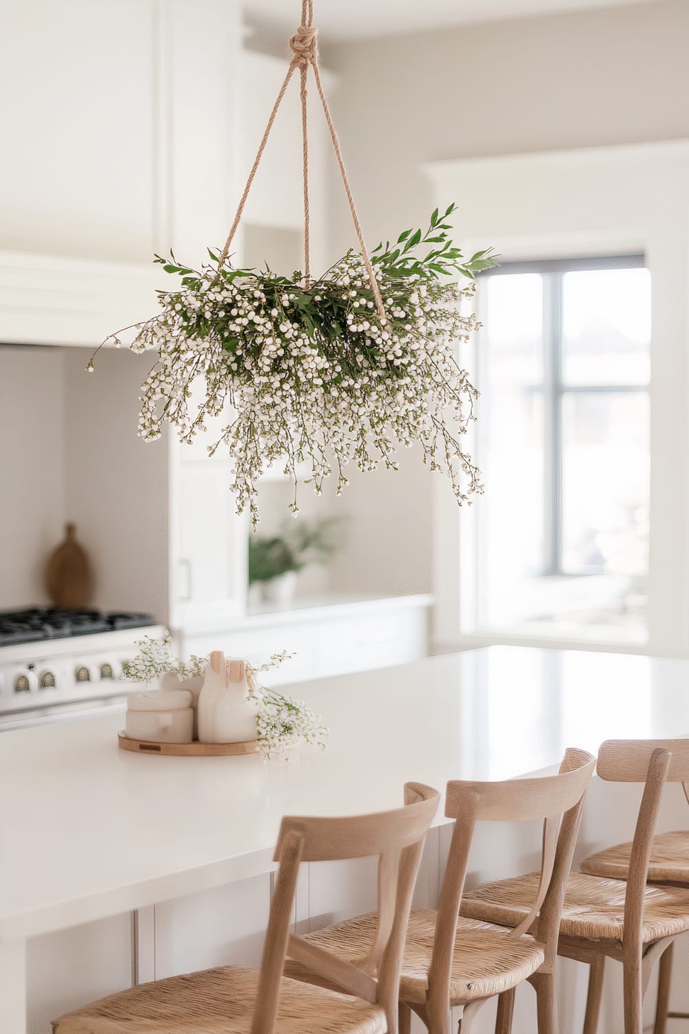 A Scandinavian-inspired kitchen featuring a simple hanging mistletoe arrangement above a white kitchen island. The mistletoe is adorned with small white berries and green leaves. The island is surrounded by wooden chairs with woven seats, and there is a small tray with white decor items and greenery on the island. The kitchen is bright with natural sunlight coming through a large window in the background.