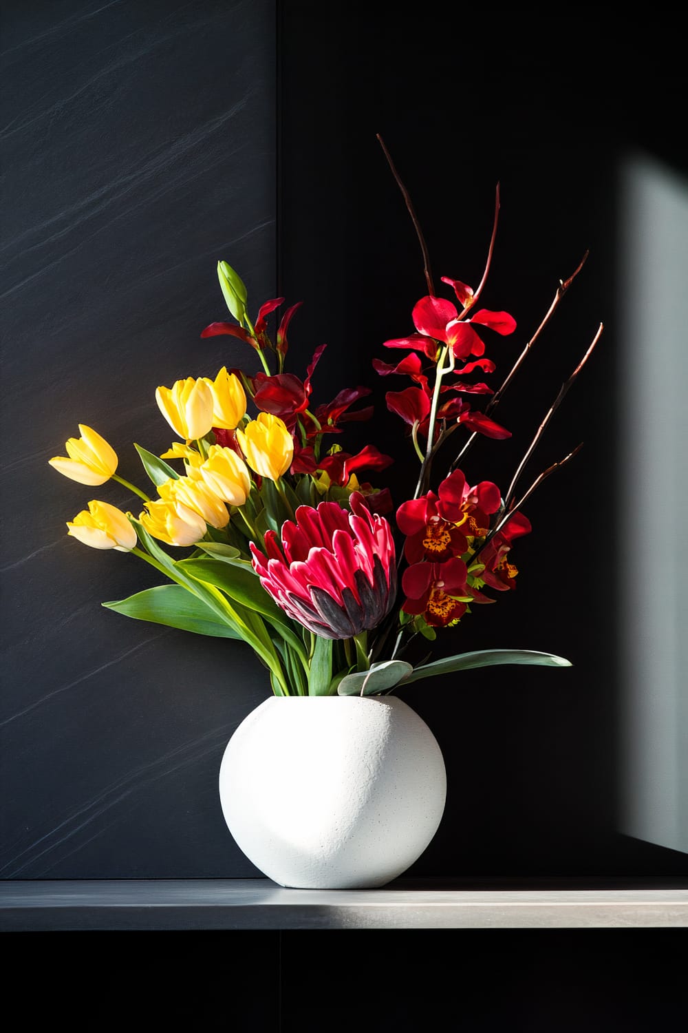 A modern, minimalist vase with a smooth, round surface is filled with a vibrant arrangement of flowers. The bouquet features bright yellow tulips, deep red orchids, and a striking pink protea. The vase is placed on a sleek, dark surface against a dark background, with sunlight casting a soft shadow.