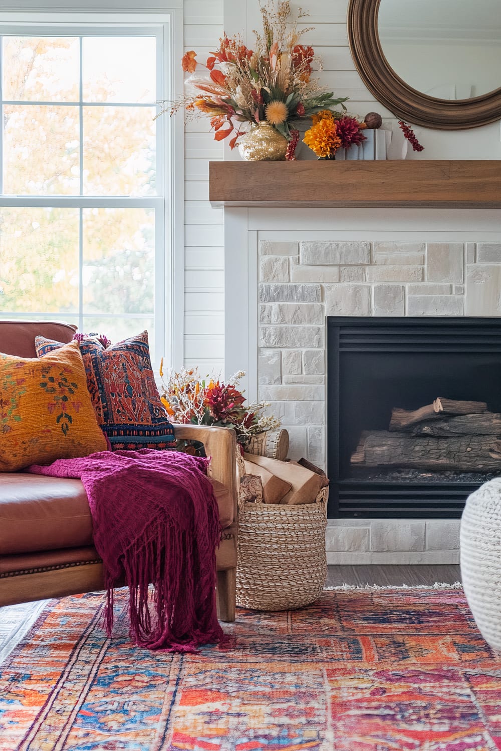 A living room with a white textured brick fireplace and a wooden mantel. A round mirror hangs above the mantel, and a decorative vase filled with vibrant autumn foliage sits on top. To the left, a brown leather armchair is adorned with colorful throw pillows and a deep magenta knitted throw blanket. A wicker basket containing firewood sits beside the fireplace. The room’s floor is covered with a multicolored, patterned rug.