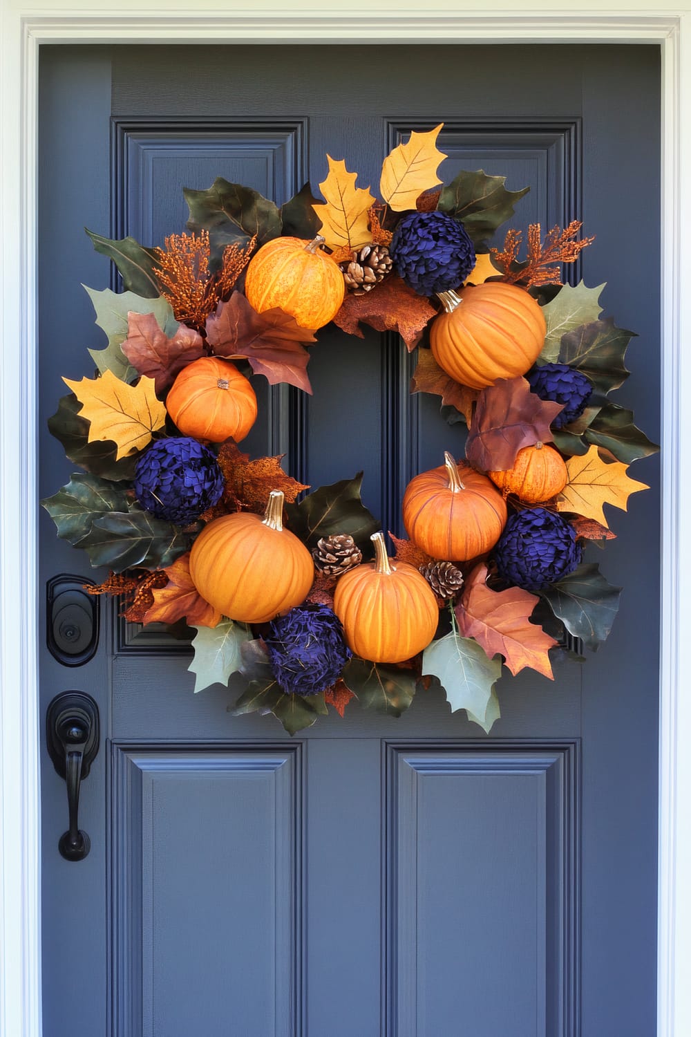 A fall-themed wreath hanging on a dark blue door. The wreath is adorned with small orange pumpkins, pine cones, deep purple flowers, and autumn leaves in shades of yellow, red, and green.