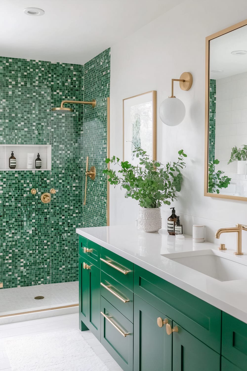 A bathroom interior featuring green mosaic tiles on the shower wall, extending to a section of the main wall. The shower area includes gold fixtures, such as a rainfall showerhead and a handheld shower. To the right, there is a sink with a white countertop and green cabinets, adorned with gold handles and hardware. Above the sink is a large mirror with a gold frame, and beside it, a modern wall sconce with a gold base and a round white glass shade. A potted plant in a white decorative vase sits on the countertop.