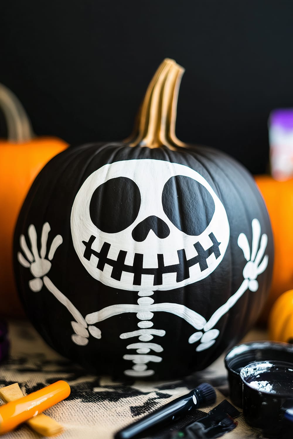 An intricately painted black pumpkin with a white skeleton design featuring large eyes, a skeletal smile, and outstretched arms. The pumpkin is surrounded by painting supplies and other pumpkins in the background.