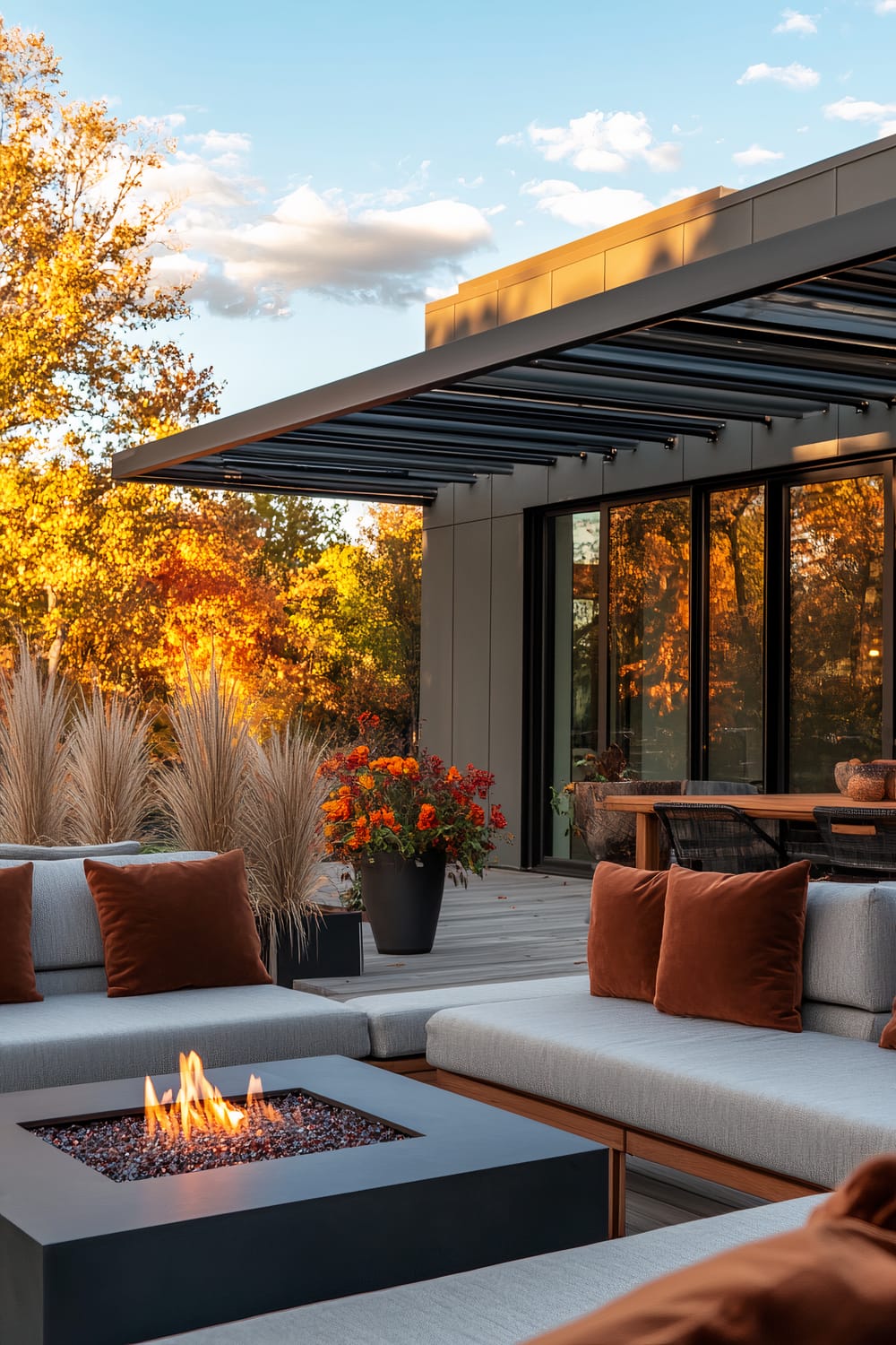 An outdoor seating area with modern decor against an autumn backdrop. The patio features a sleek metallic pergola and glass doors. Comfortable gray sofas with burnt orange cushions surround a square fire pit. Potted plants with vibrant foliage and tall ornamental grasses enhance the setting.