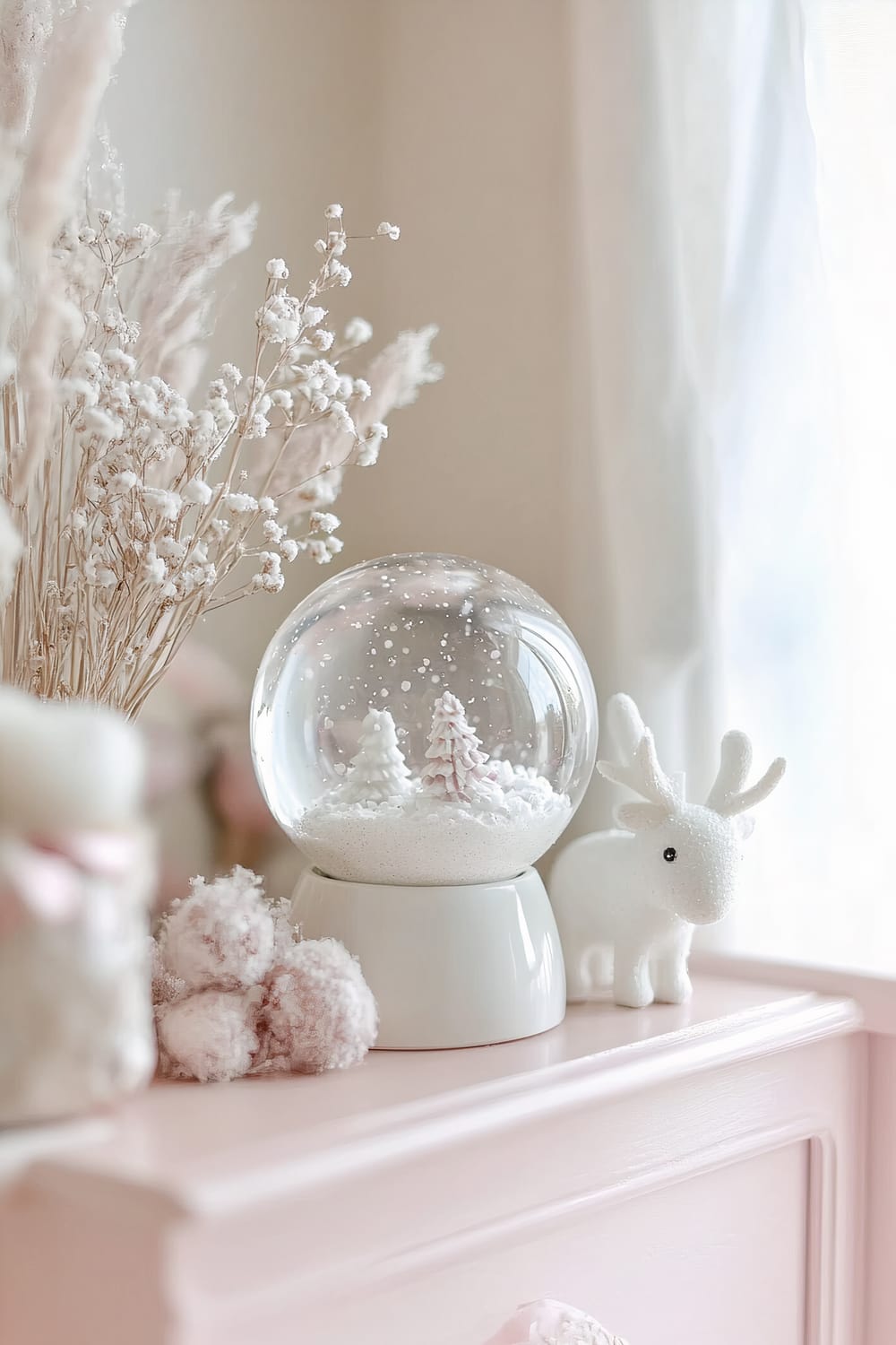 A white ceramic snow globe containing miniature snowy trees is placed on a pastel pink shelf in a nursery. There is also a white reindeer figurine nearby, and dried flowers in the background, all softly illuminated by natural daylight.