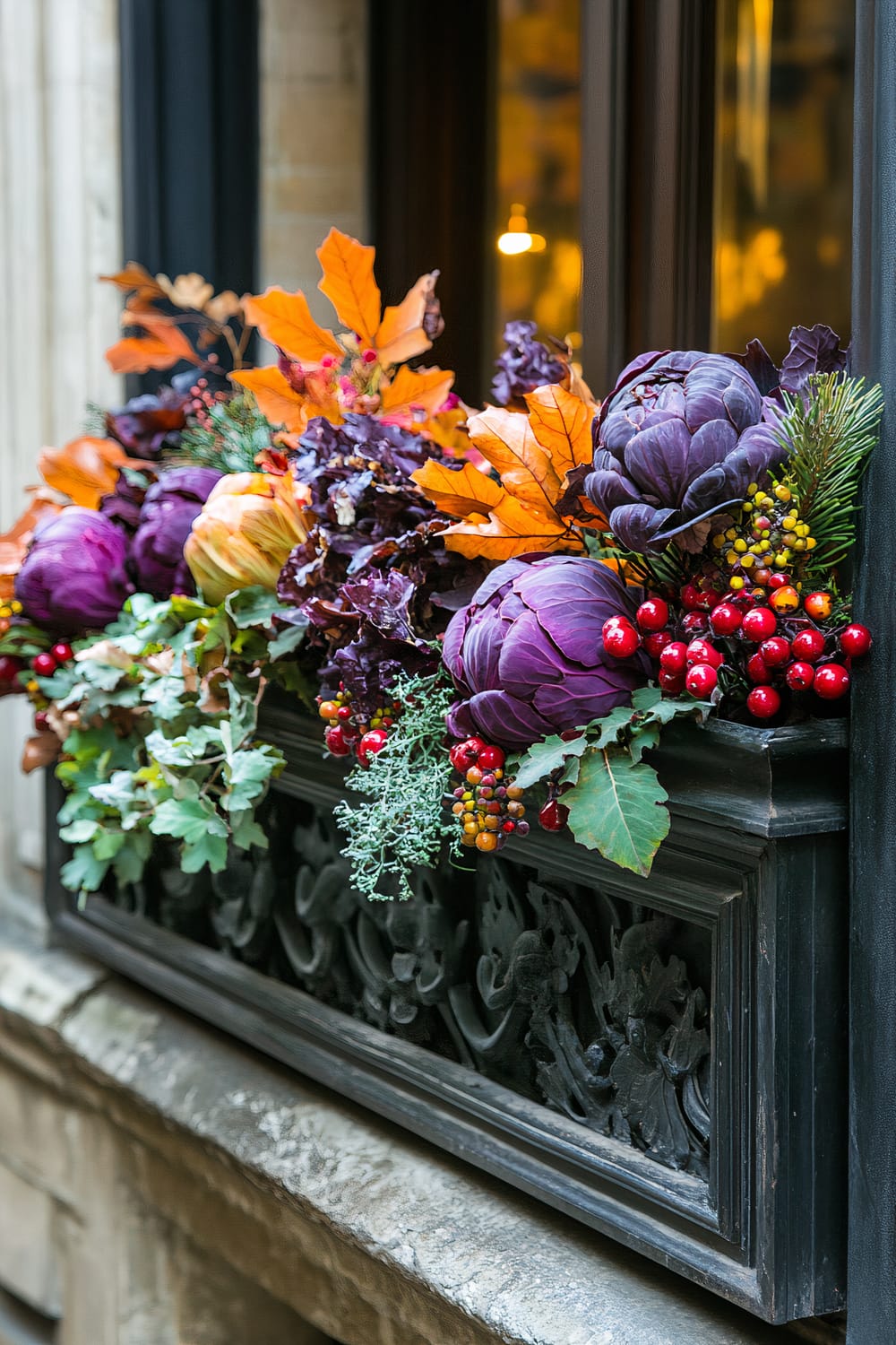 A window box filled with a vibrant autumn-themed arrangement. The arrangement includes deep purple ornamental cabbages, bright orange leaves, red berries, and green foliage. The black ornate window box provides a striking contrast to the colorful display. The background shows a softly lit window and part of a building facade.