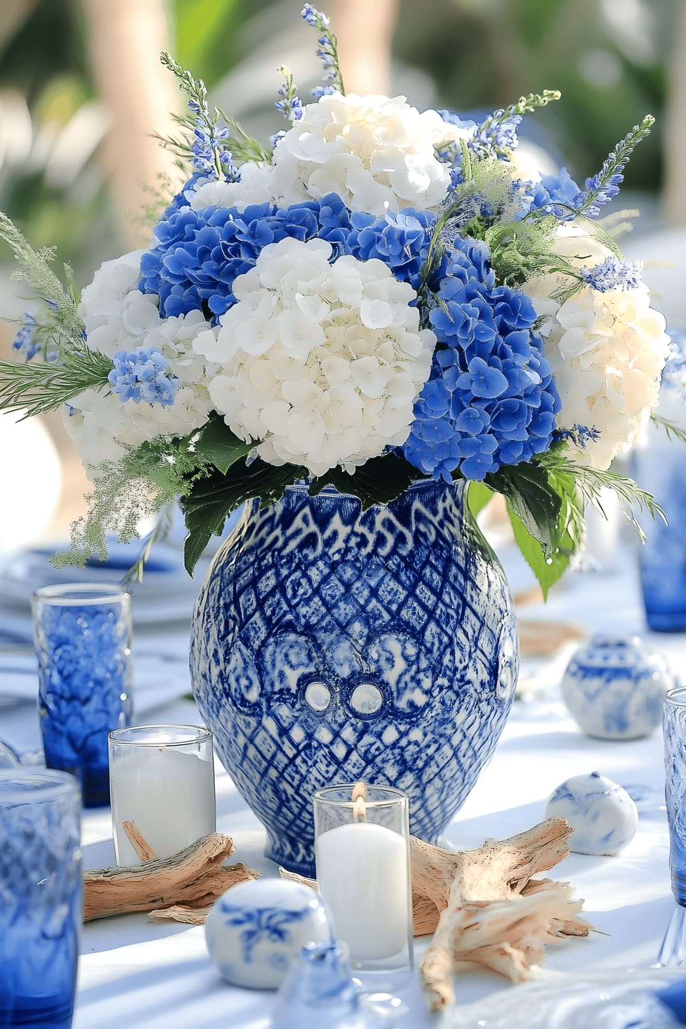 A large, ornate blue ceramic vase filled with white and blue hydrangeas and delphiniums, set as a centerpiece on a table covered with a white tablecloth. The vase is surrounded by white candles in blue glass holders and small pieces of driftwood. The table is set with blue-and-white patterned plates. The scene is bathed in bright, natural sunlight. The clean, coastal colors and stunning blue-and-white contrast evoke the serene beauty of Santorini.