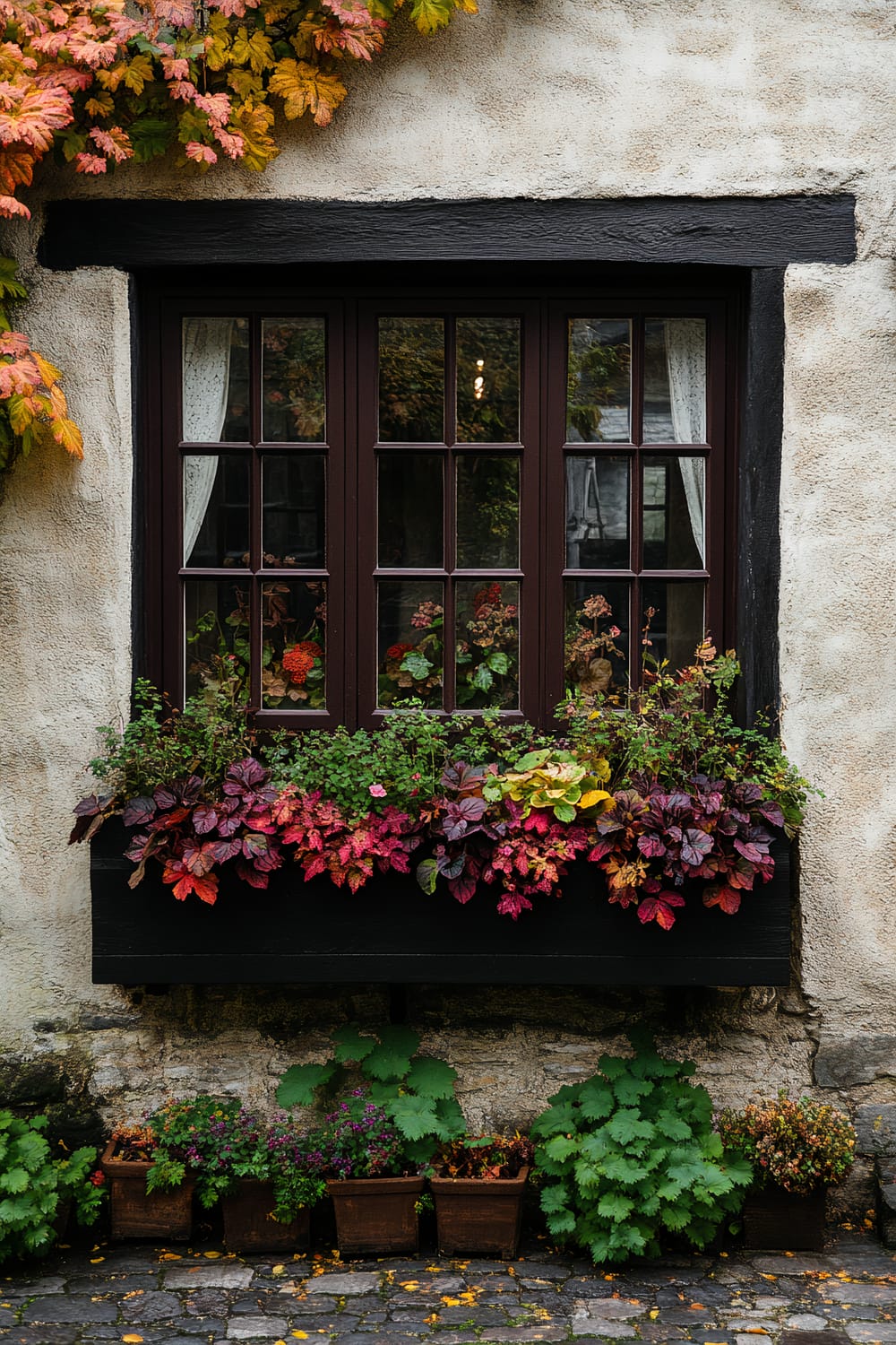 A charming window with wooden frames in a quaint stone wall adorned with colorful autumn leaves. Below the window, a planter brims with a variety of vibrant flowers in shades of red, purple, orange, and green. Several pots with more greenery and flowers are placed along the base of the wall, complementing the overall look.