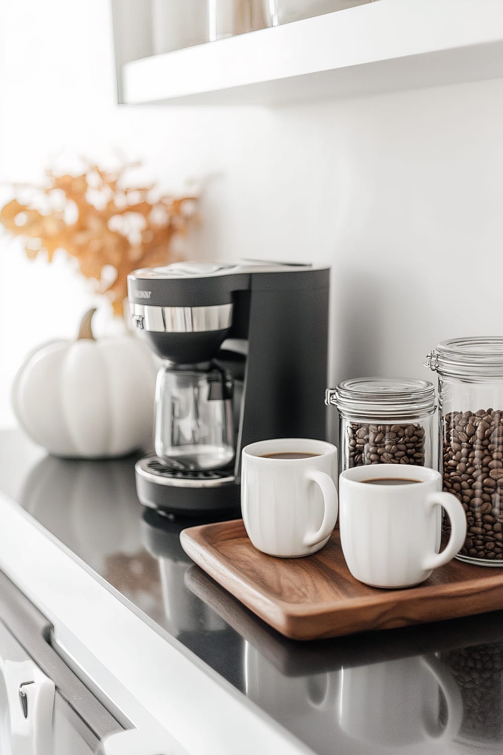 A neatly arranged coffee station featuring a modern black coffee maker, two mugs filled with coffee on a wooden tray, and two jars of coffee beans. The countertop also contains a decorative white pumpkin and dried autumn leaves in a white vase.