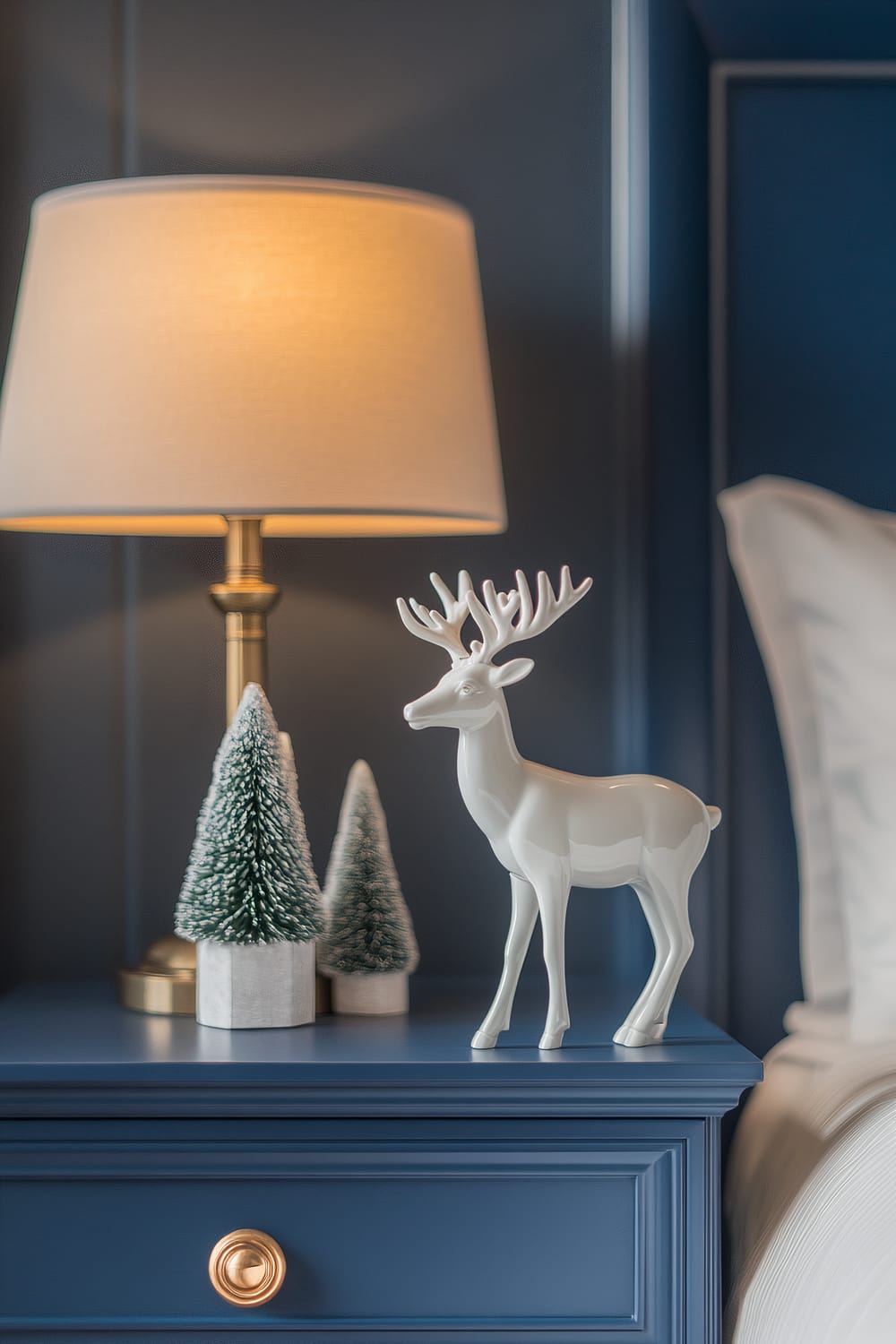 A white porcelain reindeer figurine is placed on a cobalt blue nightstand in a bedroom. The nightstand also features two small, green, snow-covered miniature trees. To the left of the reindeer, there is a brass bedside lamp with a beige lampshade that emits a warm, soft light. The edge of a white bed pillow is visible on the right side of the nightstand.