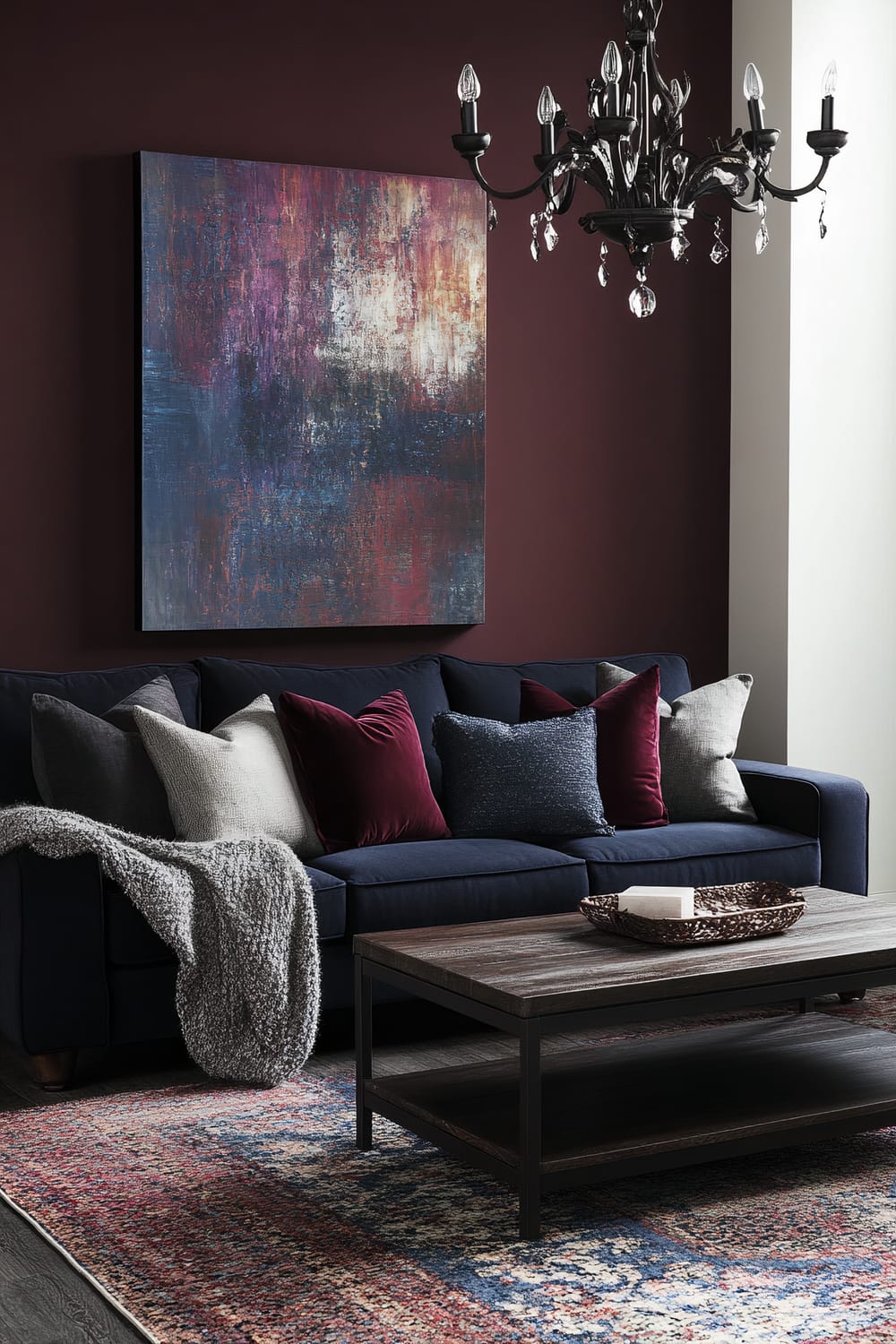 A sophisticated living room with moody hues, featuring a plush navy sofa with burgundy and gray throw pillows, a large dark wood coffee table, and a wool throw. Ambient light from a stylish chandelier enhances the rich color palette. An abstract art piece decorates the wall.