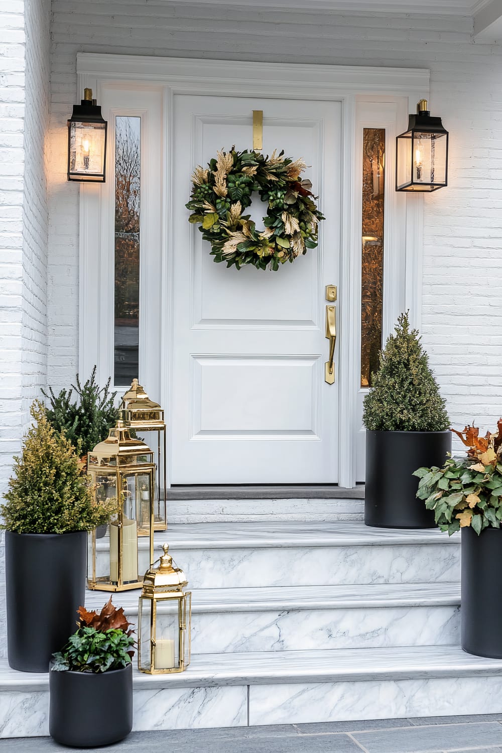 A front door decorated for the fall season. The white door is flanked by tall windows and lit by black lantern-style sconces. A festive wreath hangs on the door, and several black planters with greenery and ornamental plants line the steps, accompanied by gold lanterns with candles.