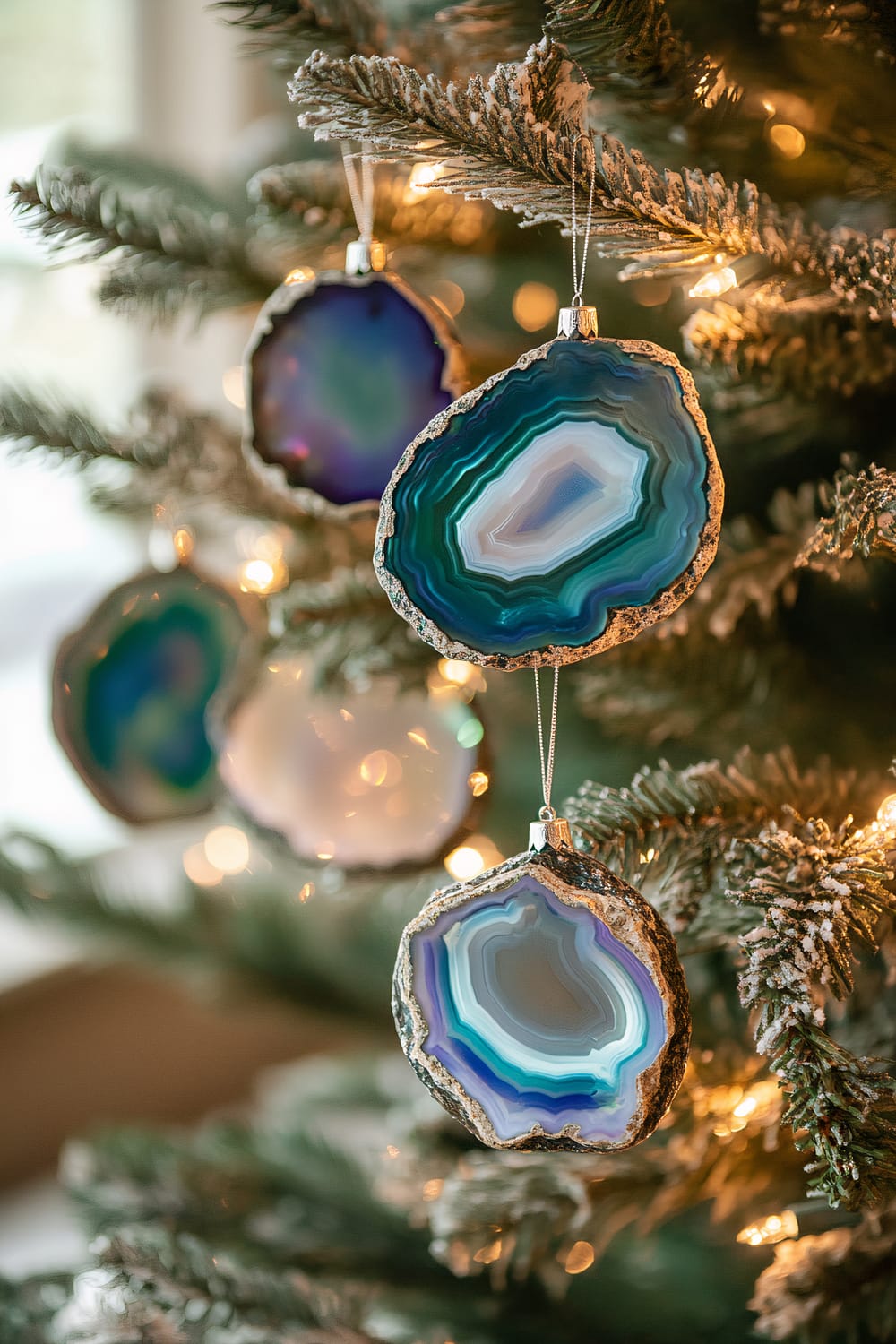 Close-up view of Christmas tree ornaments, featuring agate geode slices in varying shades of blue, green, and purple. The ornaments are hung on a frosted fir tree with fairy lights.