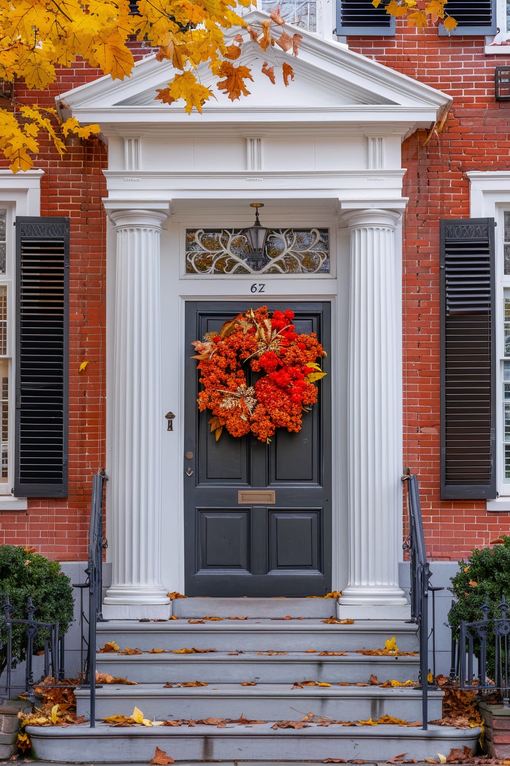 An exterior view of a house entrance is seen with brick walls and classic architectural elements. The focal point is a dark grey front door adorned with a vibrant orange autumn wreath. The doorway is framed by white columns and a decorative lintel. The entrance steps are sprinkled with fallen autumn leaves. The house features black shutters on either side of the door and a partially visible lamp above the door.