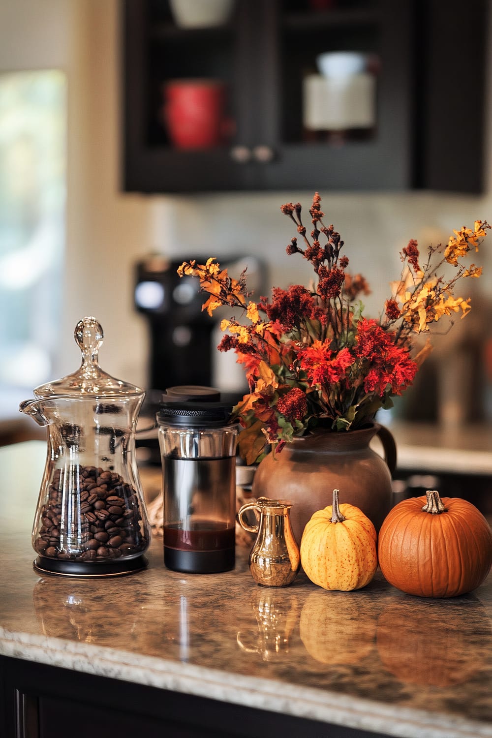 A kitchen counter with an autumn-themed display featuring a glass jar filled with coffee beans, a coffee French press, a small golden creamer, two pumpkins, and a vase with vibrant autumn flowers.