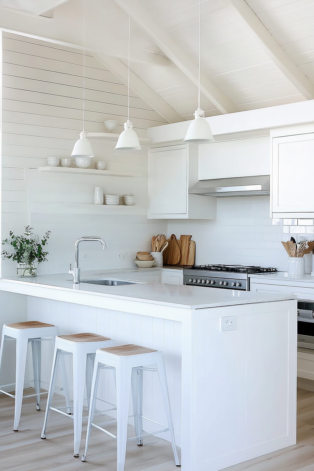 An all-white kitchen featuring a marble countertop with an integrated sink and silver faucet on a kitchen island. Three white barstools with wooden seats are arranged in front of the island. The backsplash and walls consist of white horizontal panels, and white cabinetry provides storage. There are open shelves holding white bowls and cups, while wooden cutting boards and utensils give a natural accent. Stainless steel appliances including a stove and hood are integrated into the cabinetry, and three white pendant lights hang from the ceiling above the island.