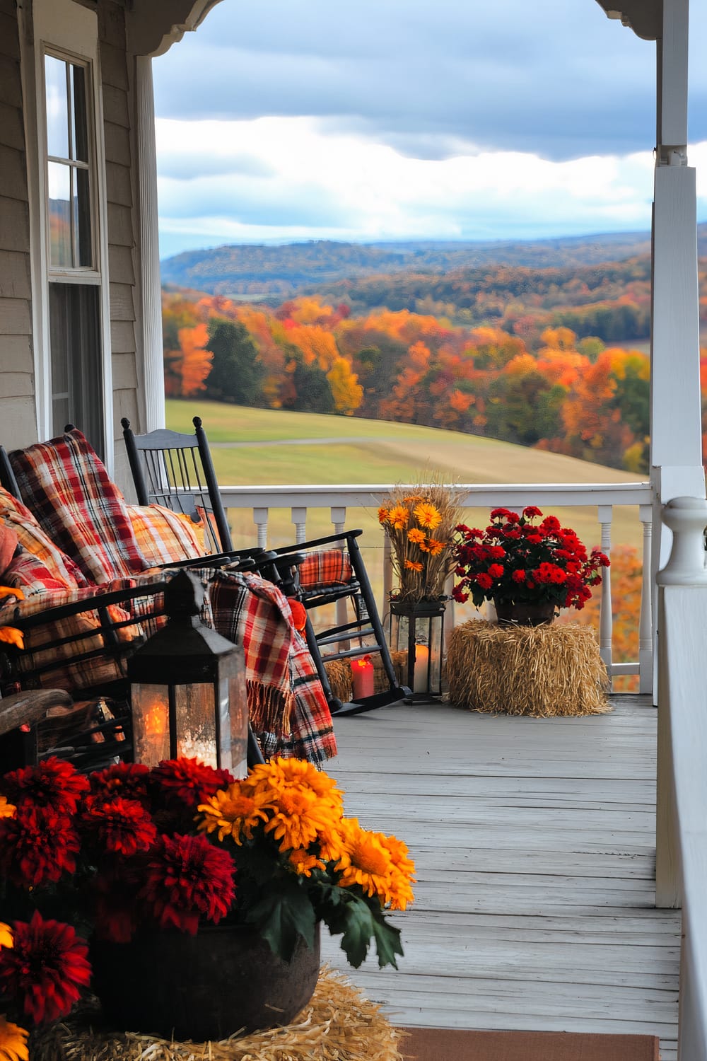 An inviting porch with a view of an autumn landscape. The porch has comfortable black rocking chairs draped with red and orange plaid blankets. There are vivid red and yellow flowers in pots placed on hay bales, a black lantern, and a rustic decor that adds to the warm setting. The background shows rolling hills covered in vibrant fall foliage under a cloudy sky.