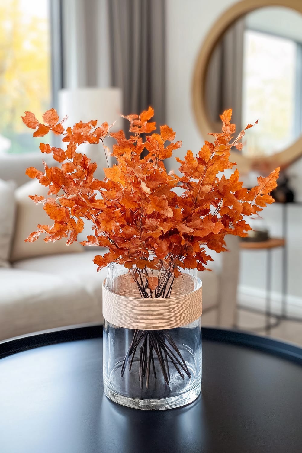 A clear glass vase filled with vibrant orange autumn leaves sits on a sleek black round table. The vase features a light beige band for added texture, and the background includes a soft, neutral-colored sofa, long grey curtains, and a round mirror adorning the wall behind.