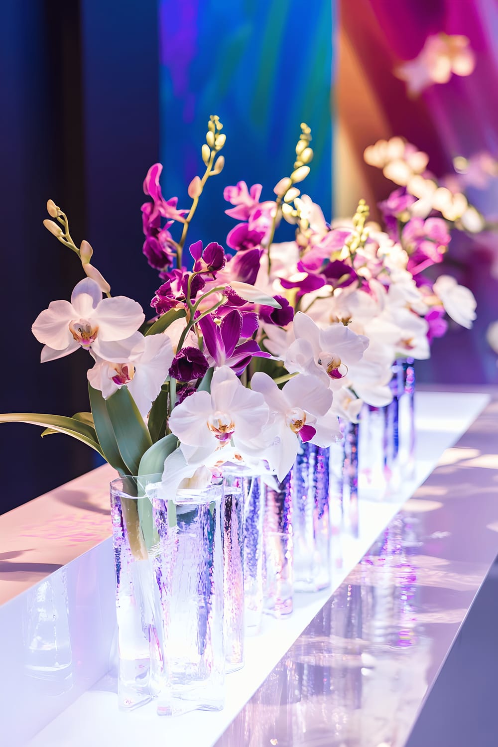 A centrepiece on a high-gloss white table featuring liquid metal flowing through transparent channels, amidst cascading fresh exotic flowers such as orchids and lilies. The backdrop consists of a vivid mural and warm LED lighting.