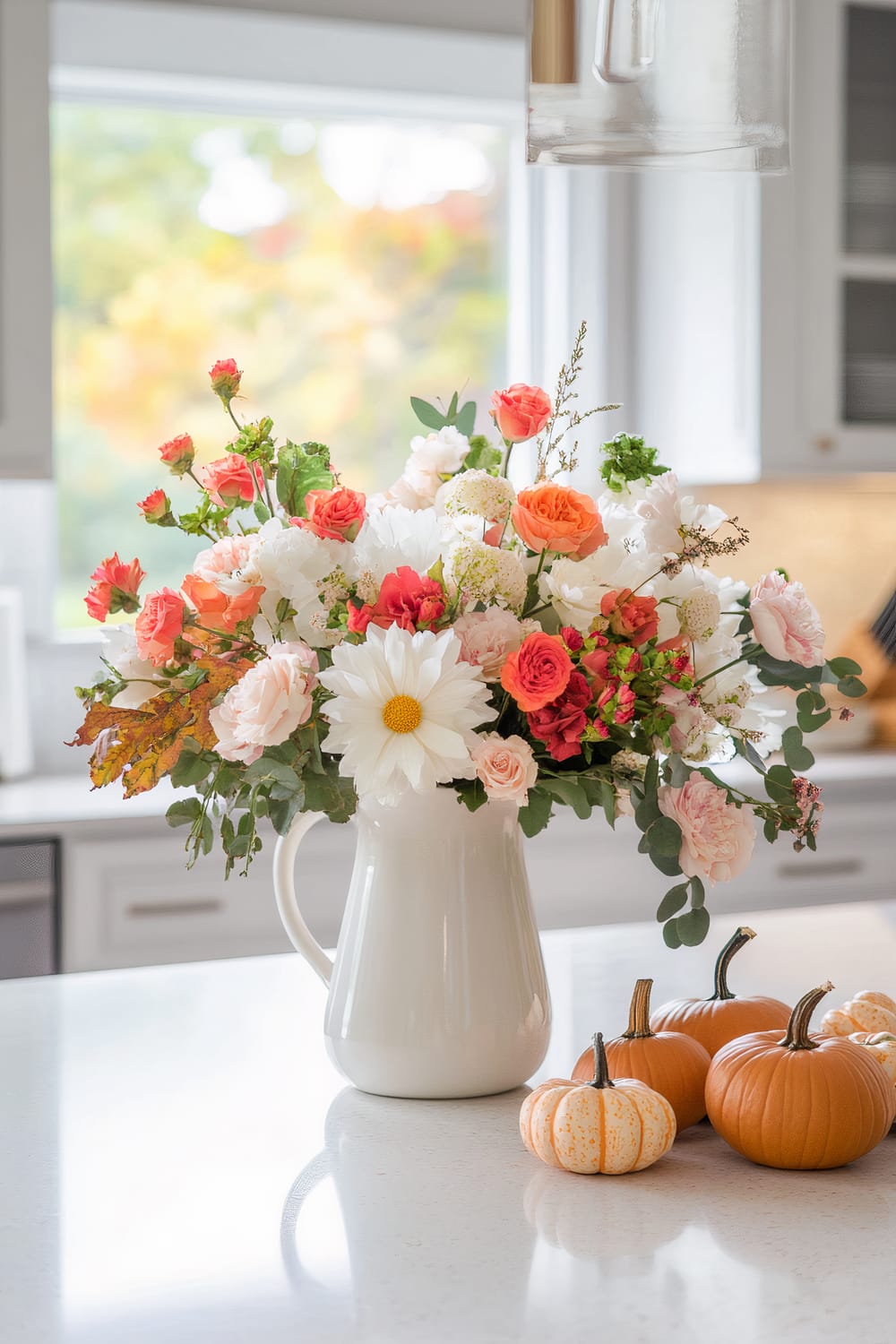 A bright and fresh floral arrangement set in a white ceramic pitcher on a white kitchen countertop. The bouquet features a variety of flowers, including white daisies, peach and pink roses, and orange blooms, complemented by lush green foliage. To the right of the pitcher are several small pumpkins in shades of orange and white, adding a seasonal autumn touch. The background shows a window with an out-of-focus view of fall foliage outside.