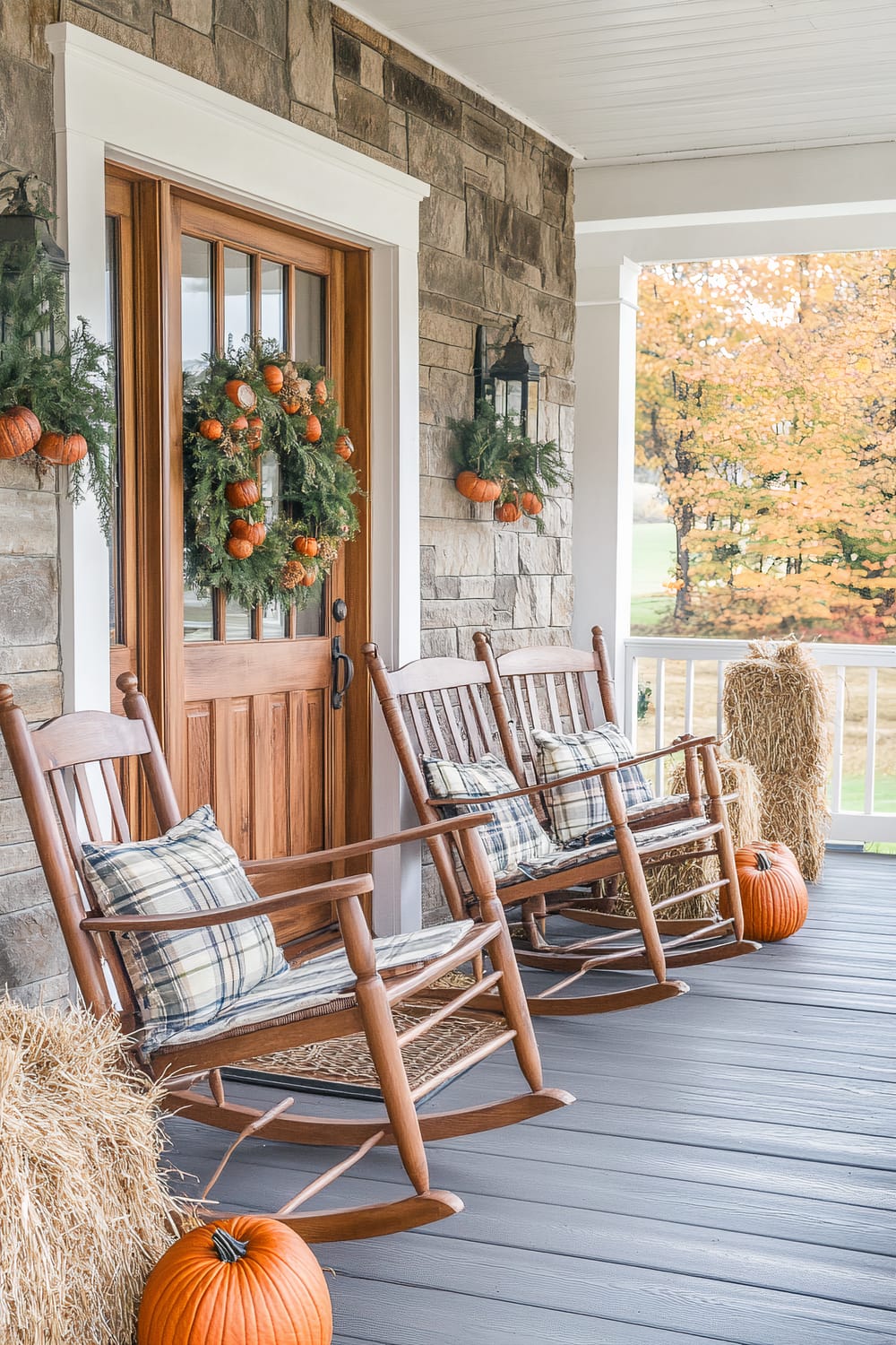 An inviting porch adorned with fall decorations, featuring a sturdy wooden front door with a large circular wreath of greenery and small orange pumpkins. Two matching wooden rocking chairs with plaid cushions are placed beside hay bales and large orange pumpkins. The porch has a wooden floor and stone walls, with black lantern-style wall lights adding to the rustic charm.