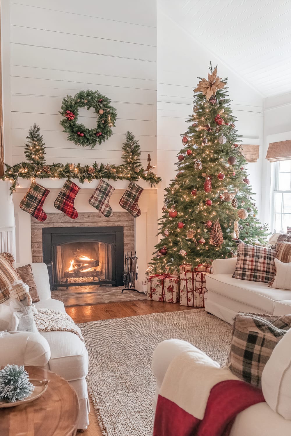 A beautifully decorated Christmas living room features a large Christmas tree adorned with ornaments and lights, several wrapped gifts placed beneath it. To the left, a fireplace with a roaring fire has a mantle decorated with garlands, small Christmas trees, and hanging plaid stockings. A wreath hangs above the mantle. The room has white walls, wood flooring, and a cozy seating area with white armchairs and couches, decorated with plaid and textured throw pillows and blankets. A wooden side table holds a small pine cone arrangement.