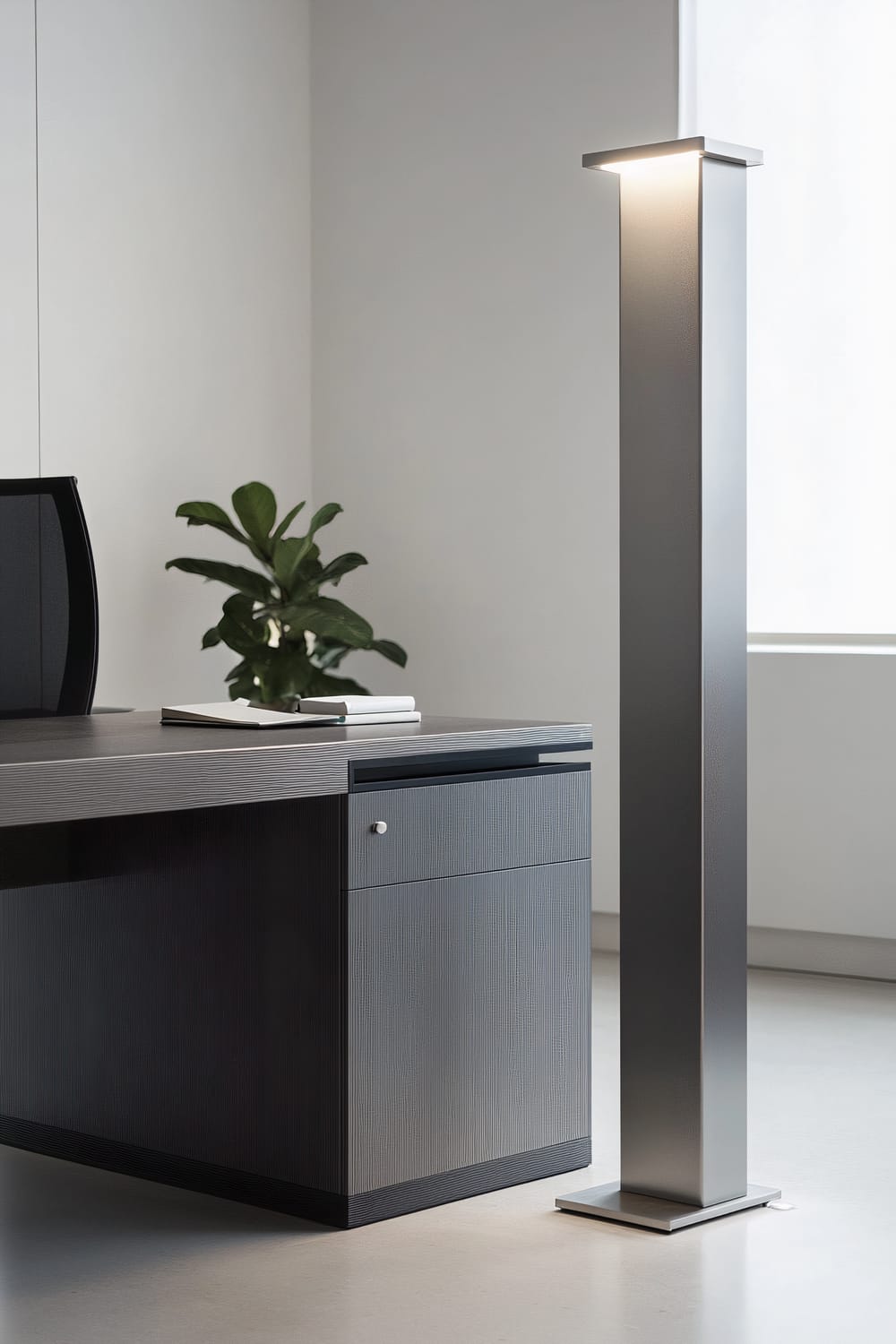 An elegant aluminum floor lamp stands next to a minimalist dark grey desk in a contemporary office space with white walls. The lamp emits a bright, focused light, illuminating the desk. A small potted plant is on the desk, adding a touch of greenery to the otherwise clean and simple decor.