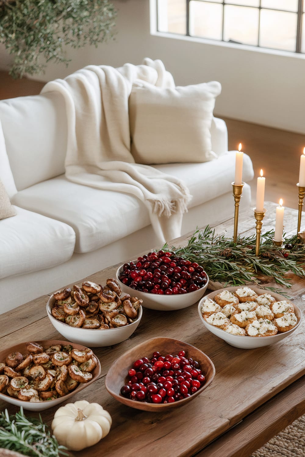 A Friendsgiving living room setup featuring a large wooden coffee table with seasonal appetizers, including stuffed mushrooms, cranberry brie bites, and mini pumpkin pies. Three ceramic bowls filled with fresh cranberries and rosemary, vintage brass candle holders with lit candles, and a chunky knit blanket on a white sofa add to the cozy setting.