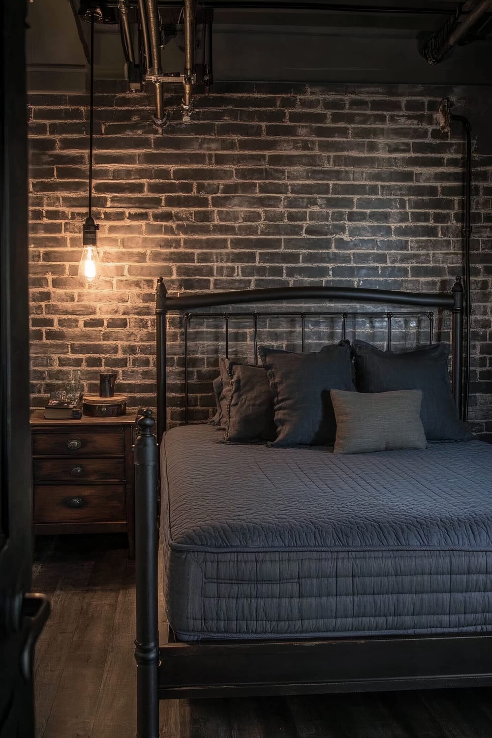 An industrial-style bedroom featuring a metal bed frame with a dark grey mattress and multiple grey pillows. An overhead Edison bulb lamp illuminates the bed. To the left of the bed is a wooden side table with a single lamp, some books, and a decorative item. The walls have a dark brick pattern, and exposed plumbing pipes are visible on the ceiling, adding to the industrial vibe.