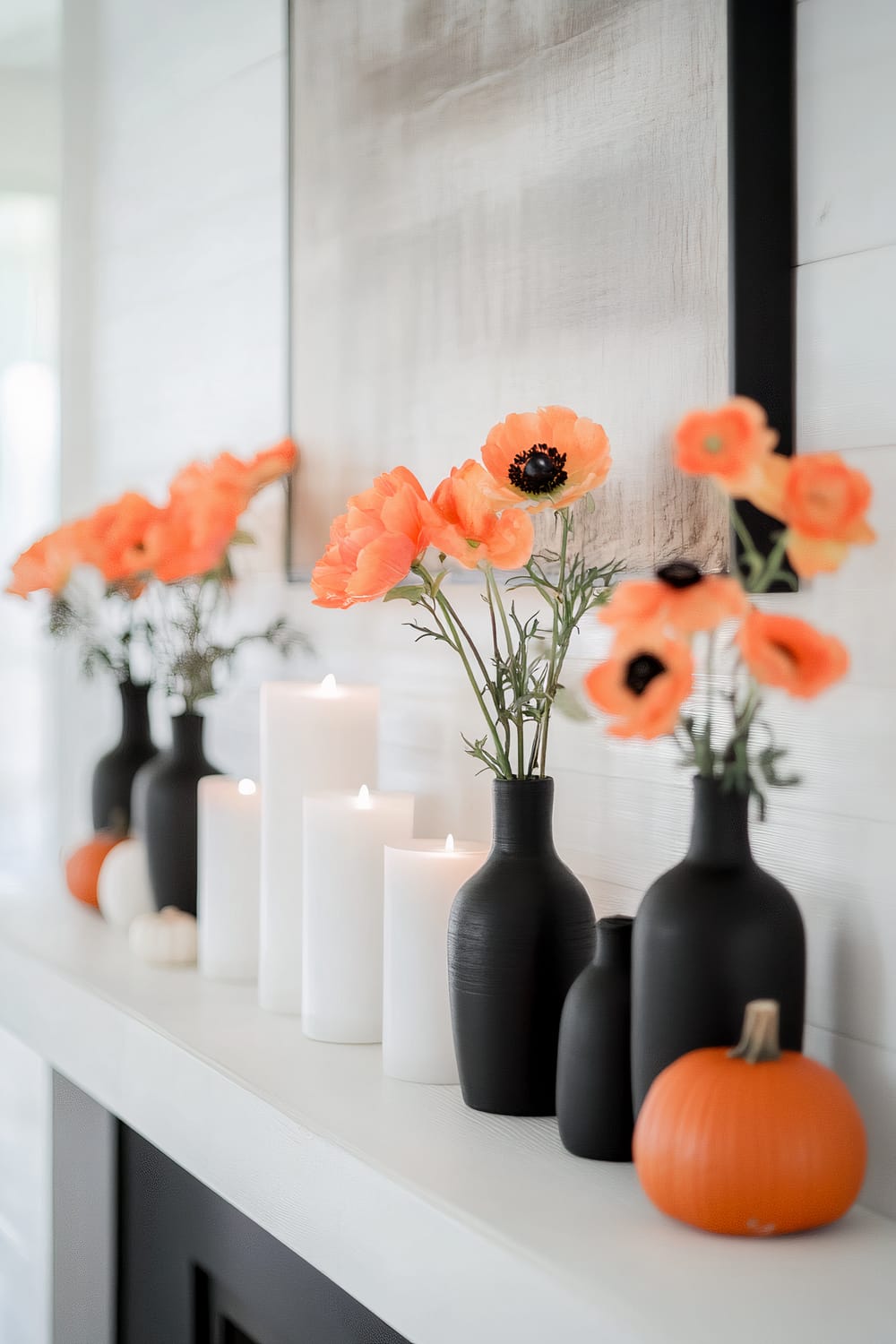 A refined mantel setting in a contemporary home, featuring an arrangement of matte black vases and vibrant orange flowers. White pillar candles of varying heights are interspersed along the mantel, casting a gentle glow. Small pumpkins are placed strategically among the decorations, adding a touch of seasonal autumn charm. The background exhibits a muted, neutral-toned wall with subtle vertical paneling, complementing the understated elegance of the display.