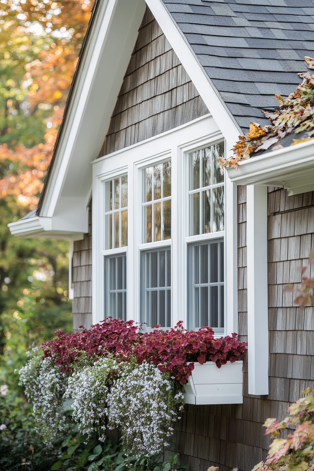 A close-up view of a home's exterior showcasing a section of the roof, shingles, and a prominent window. The window features white frames and grids, with a window box filled with vibrant red and green foliage along with delicate white flowers cascading down. The siding is made of wood shingles with a weathered grey-brown finish. The surrounding autumn foliage with hues of green and orange adds a seasonal charm to the scene.