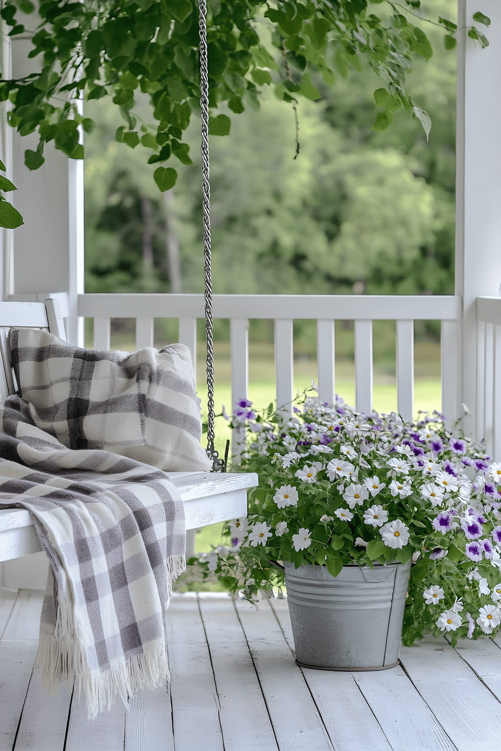 A farmhouse-inspired patio scene featuring an inviting white wooden swing bed covered in a grey and cream-toned plaid blanket. Next to the swing is a galvanized metal bucket filled with bright daisies. The flooring is made from reclaimed barn wood and the setting is completed by a charming white picket fence where morning glories in lovely shades of purple and white are climbing.