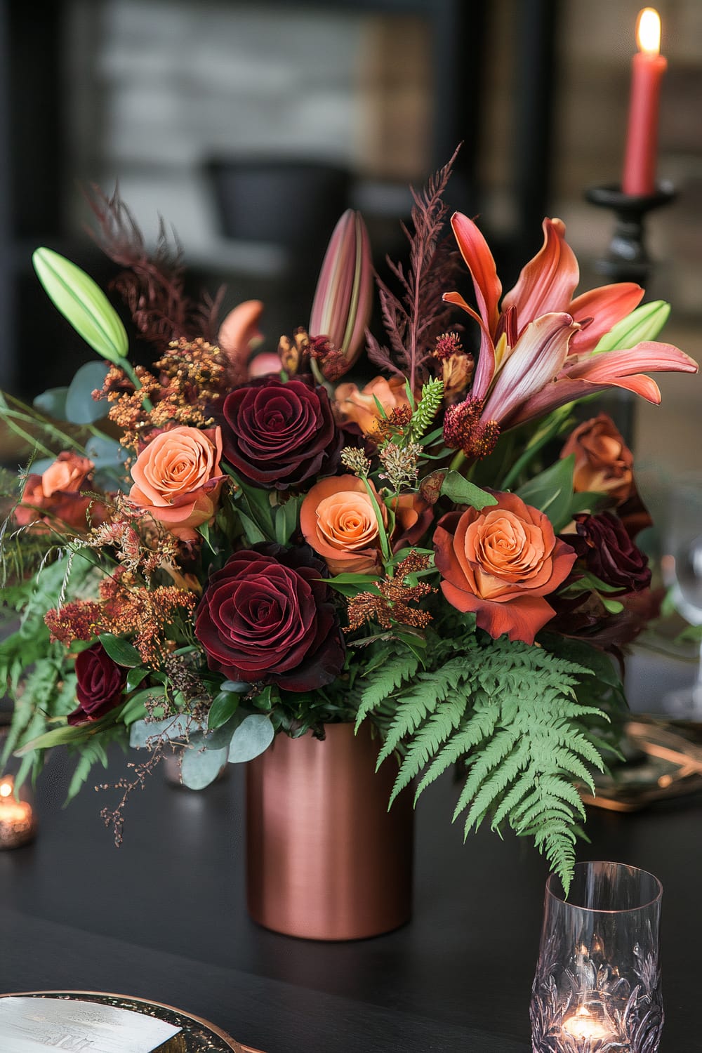 A sophisticated flower arrangement in a copper vase, featuring a mix of deep burgundy roses, peach roses, and orange lilies, complemented by fern leaves and other greenery. The arrangement is placed on a dark table beside a glass with an etched pattern and a lit red candle in the background.