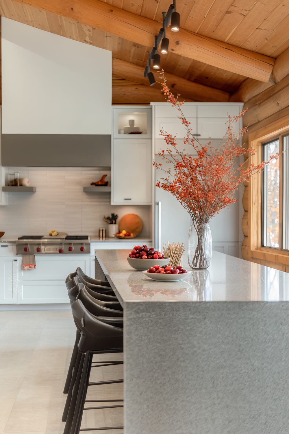 A modern kitchen with a sleek, minimalist design featuring a large central island with a polished stone countertop. Above the island, dried branches with red berries are arranged in a clear vase, adding a natural touch. The island also has plates and bowls filled with fruits. The background shows white cabinets, a stainless steel stove with red knobs, floating shelves with decor items, and a range hood. The ceiling is made of exposed wooden beams, and there is natural light coming from a window on the right side.