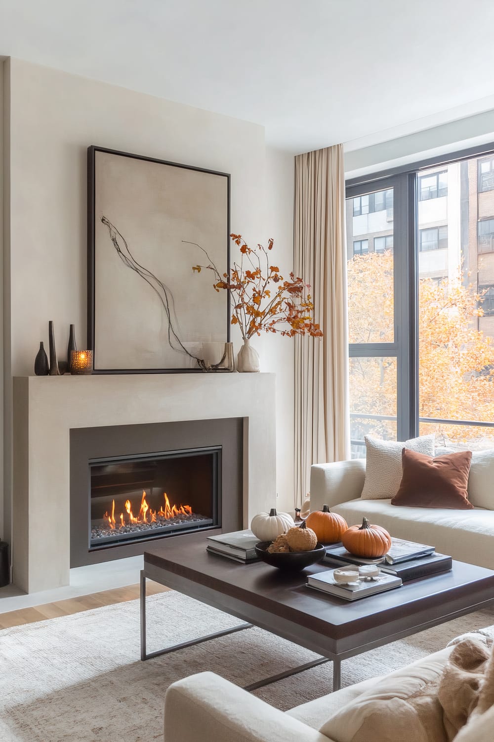 A modern living room features a clean-lined gas fireplace with soft beige tones, topped by a large abstract framed artwork and accented with decorative vases and autumnal branches. A sizable coffee table holds a collection of small pumpkins, a tray with candles, sculptural decor, and stacked books. The space is anchored by an inviting sofa with neutral and rust-colored pillows. Large windows with wall-to-wall light beige curtains offer a view of city buildings and colorful autumn foliage outside.